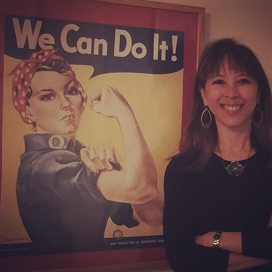“Rosie” is a photo of me at my company office standing in front of a poster of one of my “she-roes” Rosie the Riveter