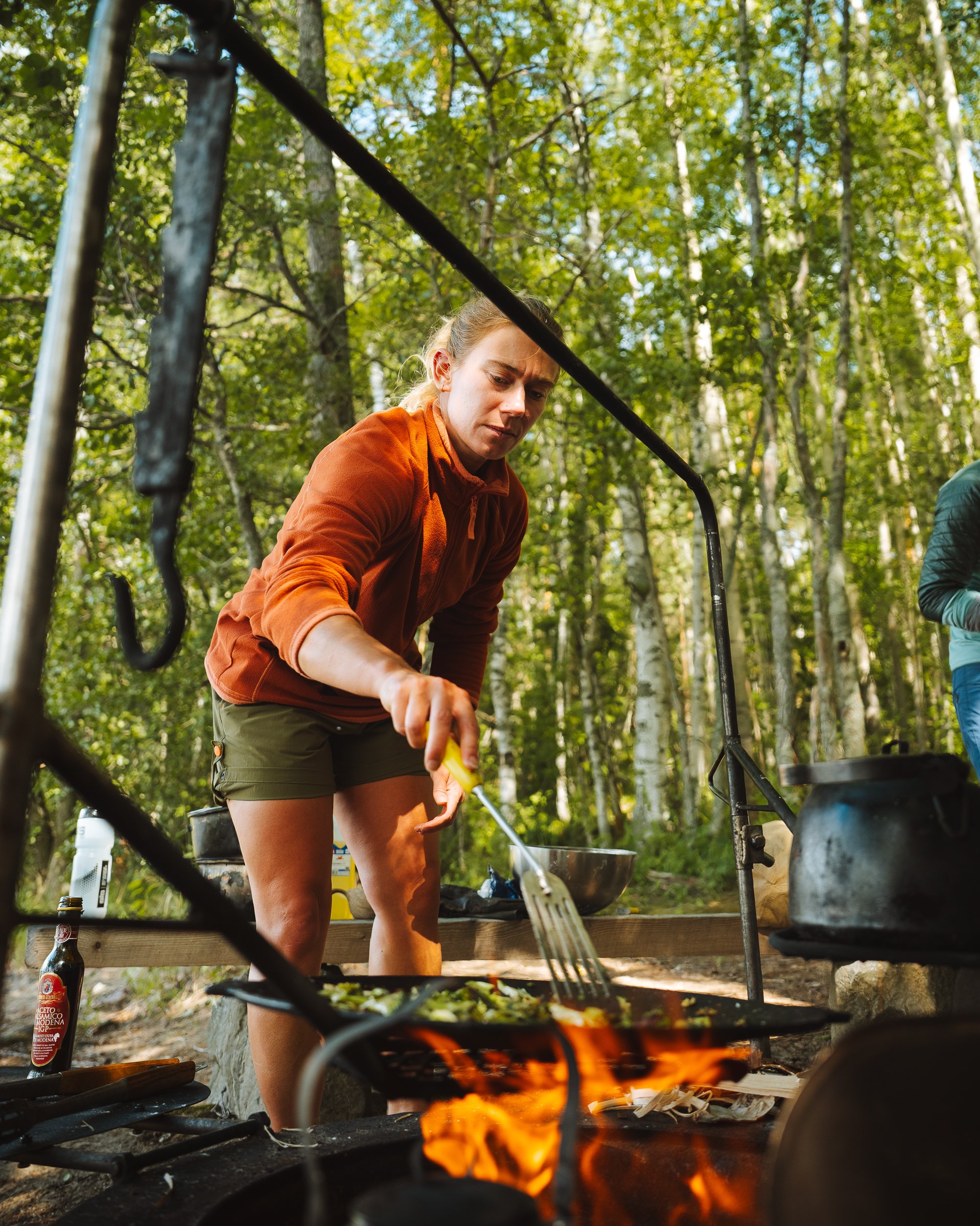 A woman in an orange jacket and green shorts cooking outdoors over a campfire, with trees and foliage in the background.