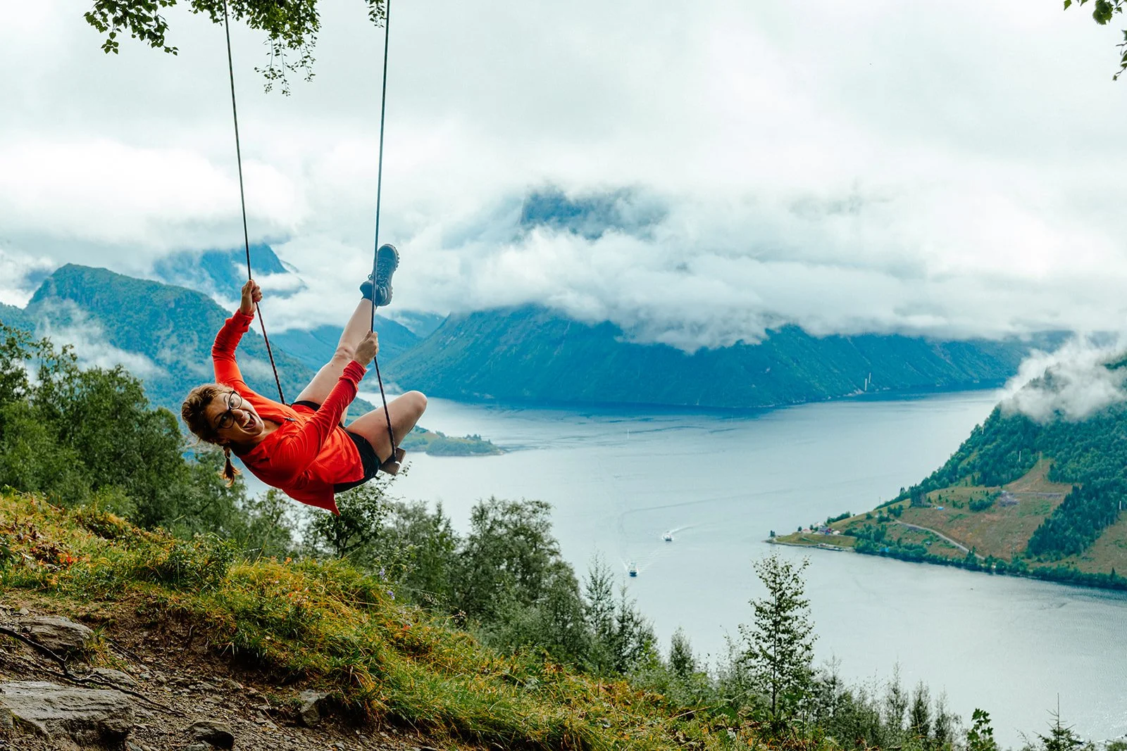A woman with glasses and a red jacket swinging on a swing set outdoors, with a large body of water, mountains, and cloudy sky in the background in Norway.