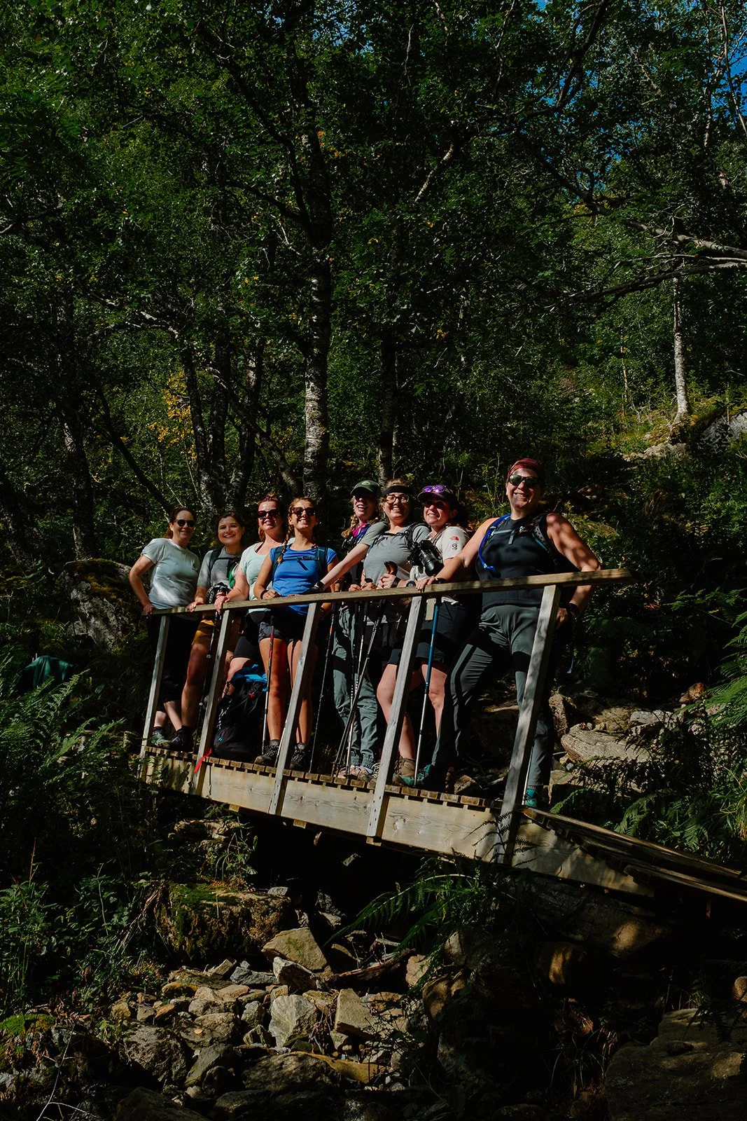A group of eight hikers standing on a wooden bridge in a forest, smiling and posing for a photo.