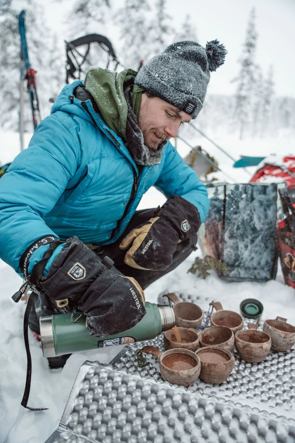 A man outdoors in snowy conditions, pouring a beverage from a thermos into clay cups on a metal tray, wearing winter clothing and gloves, with snow-covered trees in the background.