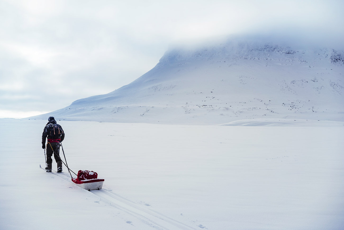 A person dressed in cold-weather gear, pulling a red sled across a snowy landscape with a large snow-covered mountain partially obscured by clouds in the background.