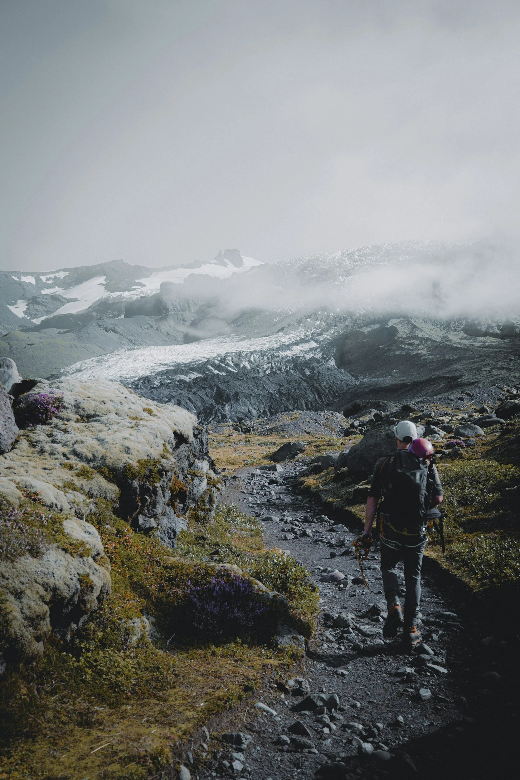 Hikers walking along a dirt trail in a mountainous, volcanic landscape with snow-capped peaks and clouds in the distance, wearing backpacks and outdoor gear.