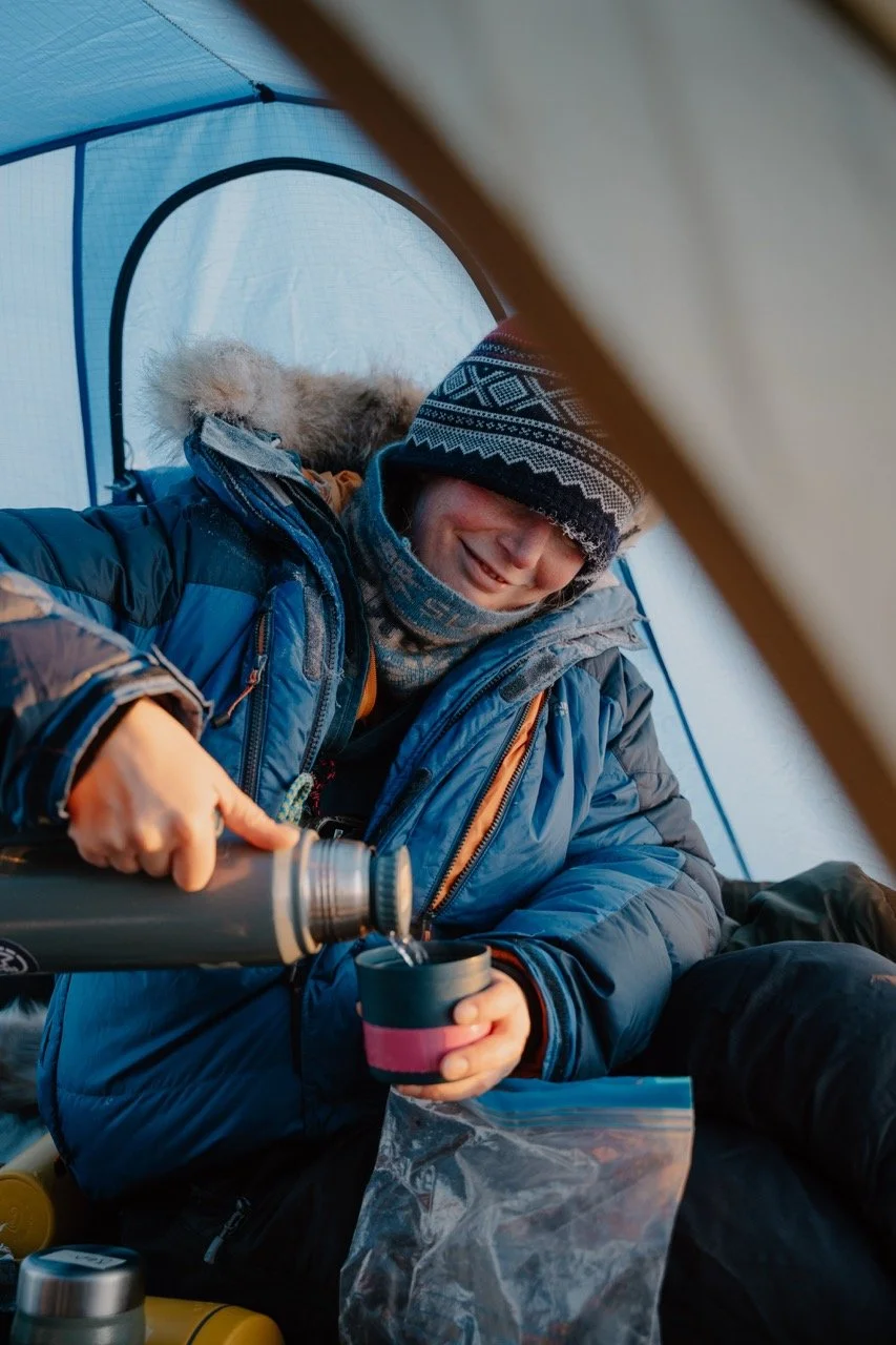Person in winter gear pouring hot beverage into a mug inside a tent.