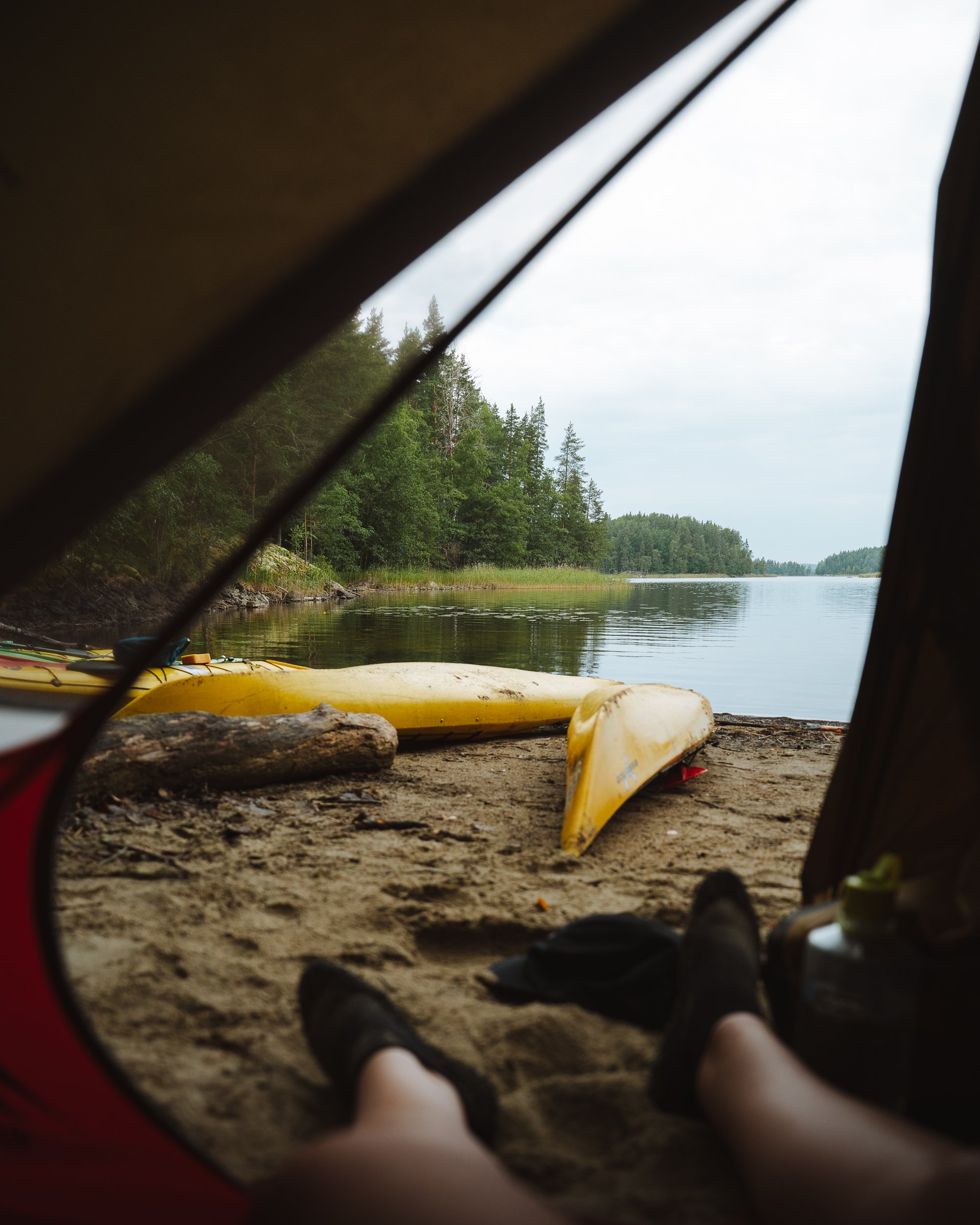 View from inside a tent looking out at a lakeside scene with trees, a sandy shore, yellow kayaks, and calm water.