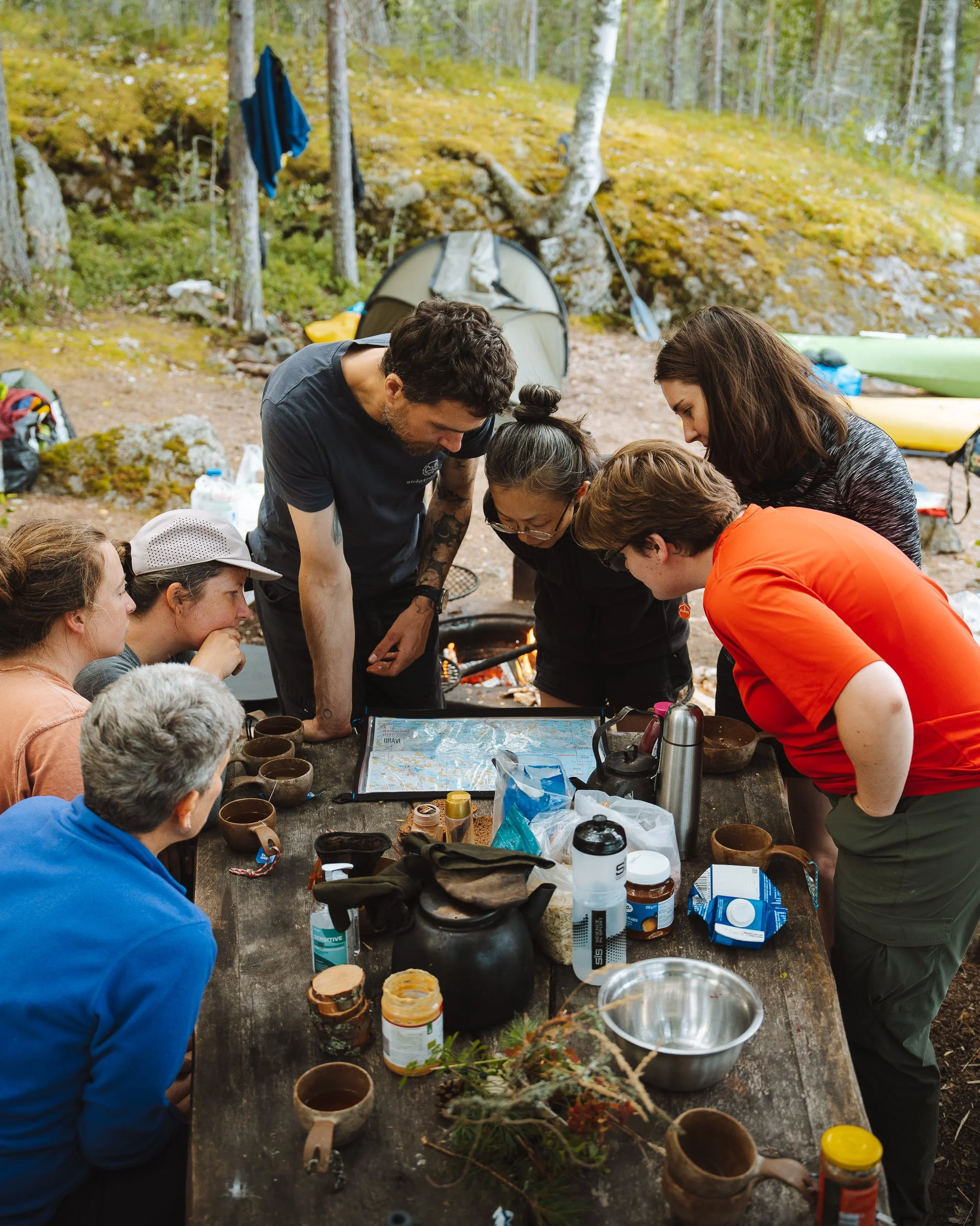 A group of people gathered around a wooden outdoor table in a forest, engaging in an activity possibly related to camping or outdoor crafts, with camping gear and supplies on the table and tents in the background.