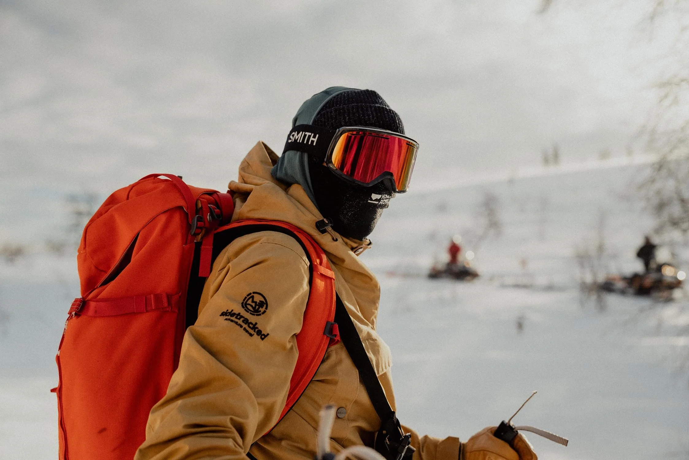 A person wearing a black beanie, red mirrored ski goggles, a beige jacket, and carrying an orange backpack, standing in a snowy landscape with snowmobiles in the background.