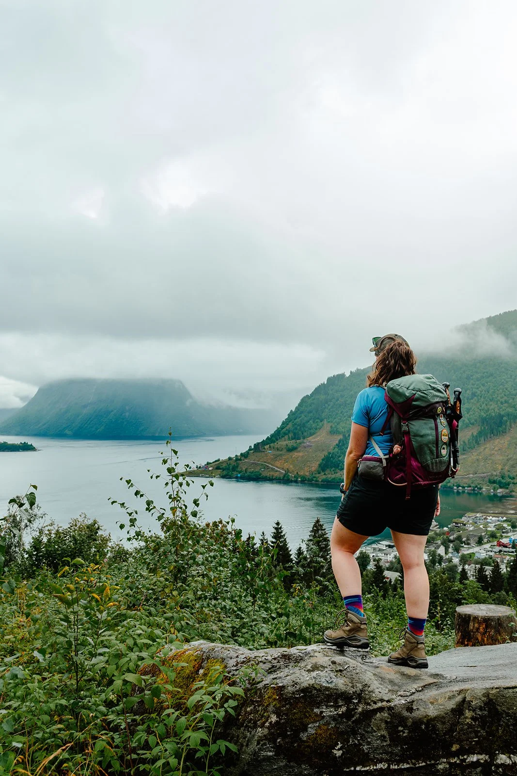 Hiker with a backpack standing on a rock, overlooking a lake surrounded by mountains under cloudy sky. Hiking in Norway.