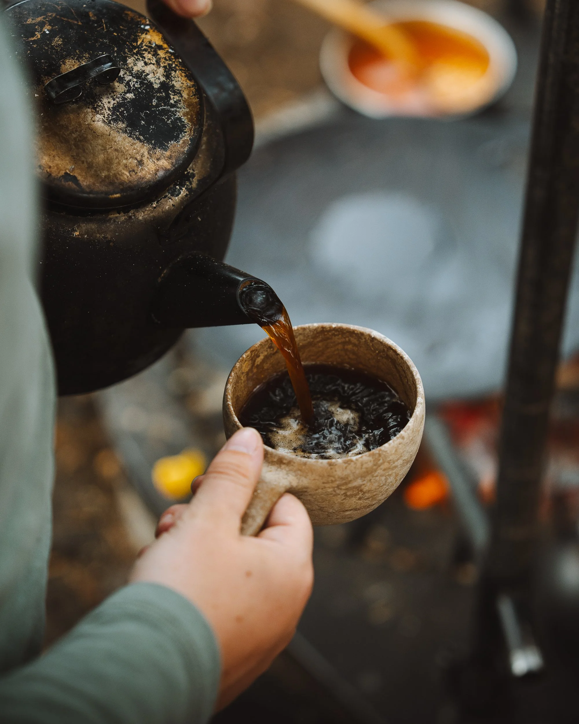 A person is pouring coffee from a coffee maker into a ceramic mug, with a cup of dark coffee already inside. The coffee maker is old and stained. The background shows a blurred kitchen or coffee shop setting.