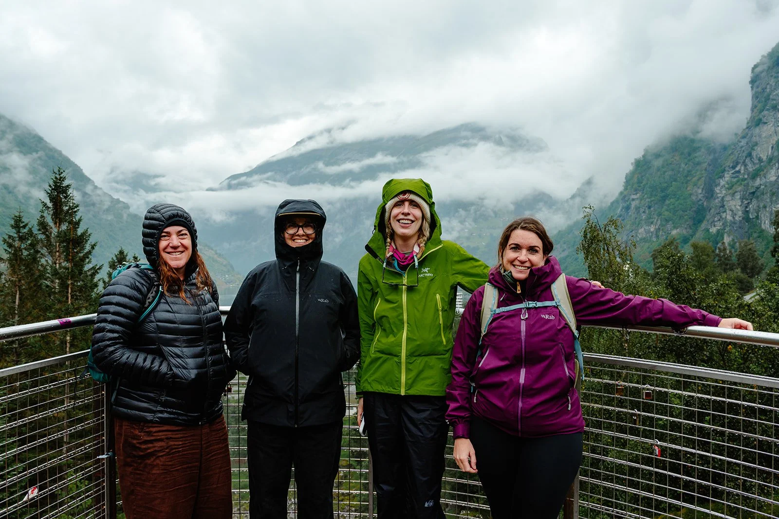 Four women standing on a balcony overlooking a misty mountainous landscape, dressed in rain jackets and outdoor gear, smiling