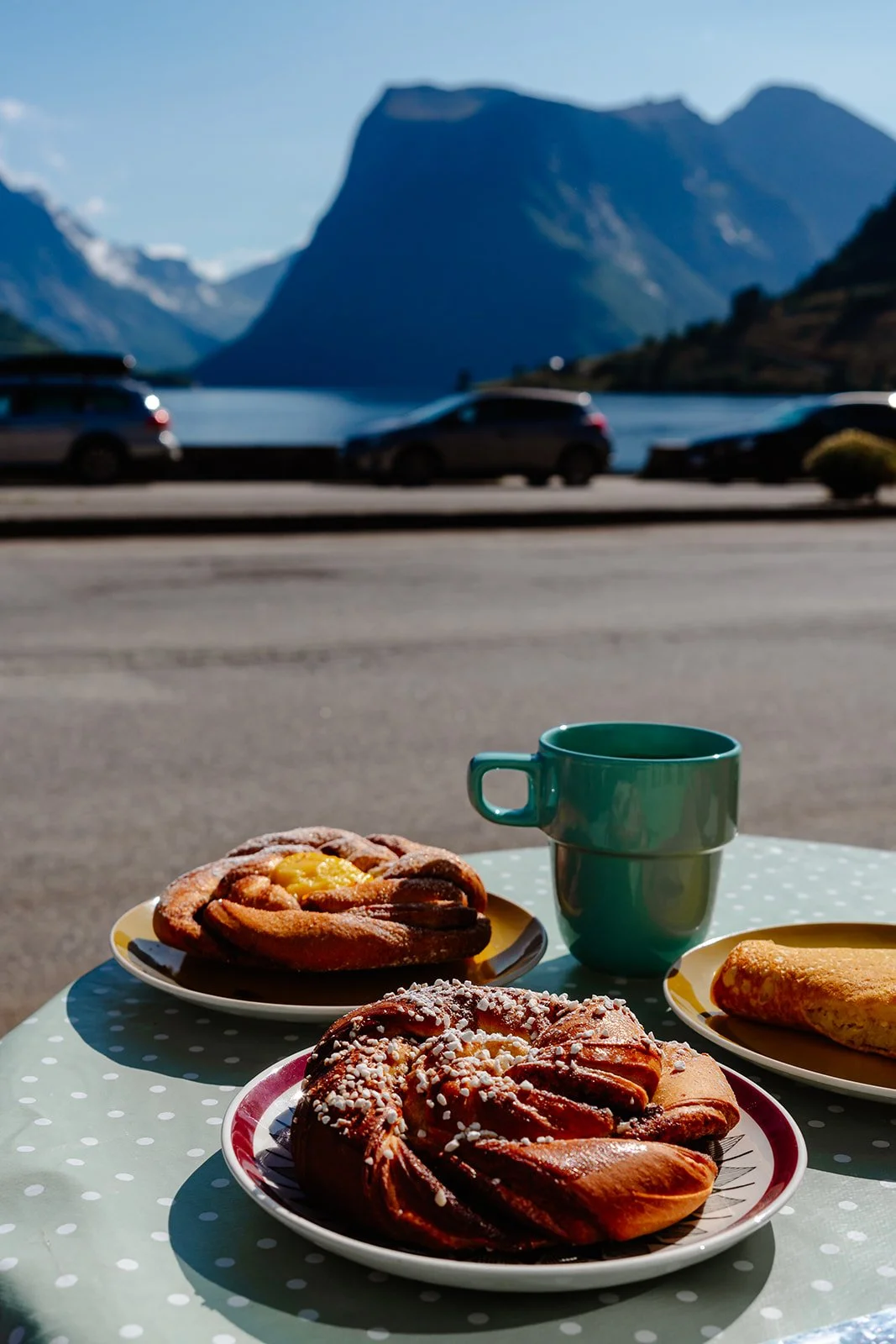 Outdoor breakfast setup with pastries and a teal mug on a polka dot tablecloth, Norwegian mountains, and lake in the background.