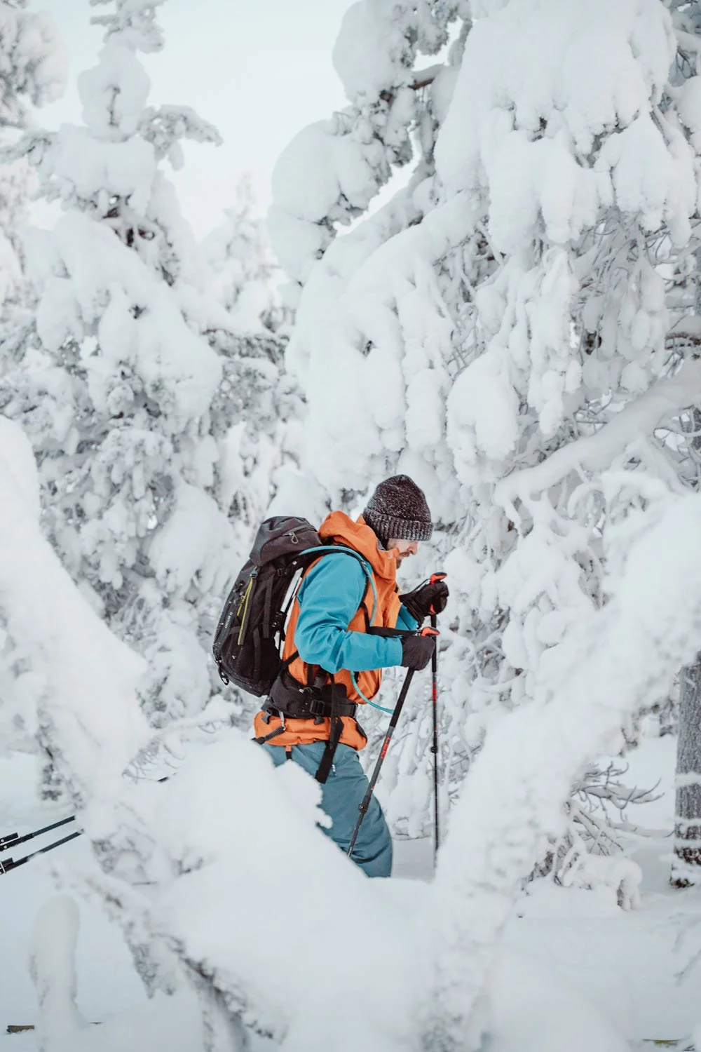 A person dressed in winter gear, including a blue jacket, orange vest, and gray beanie, stands among snow-covered trees while holding trekking poles in a snowy forest.