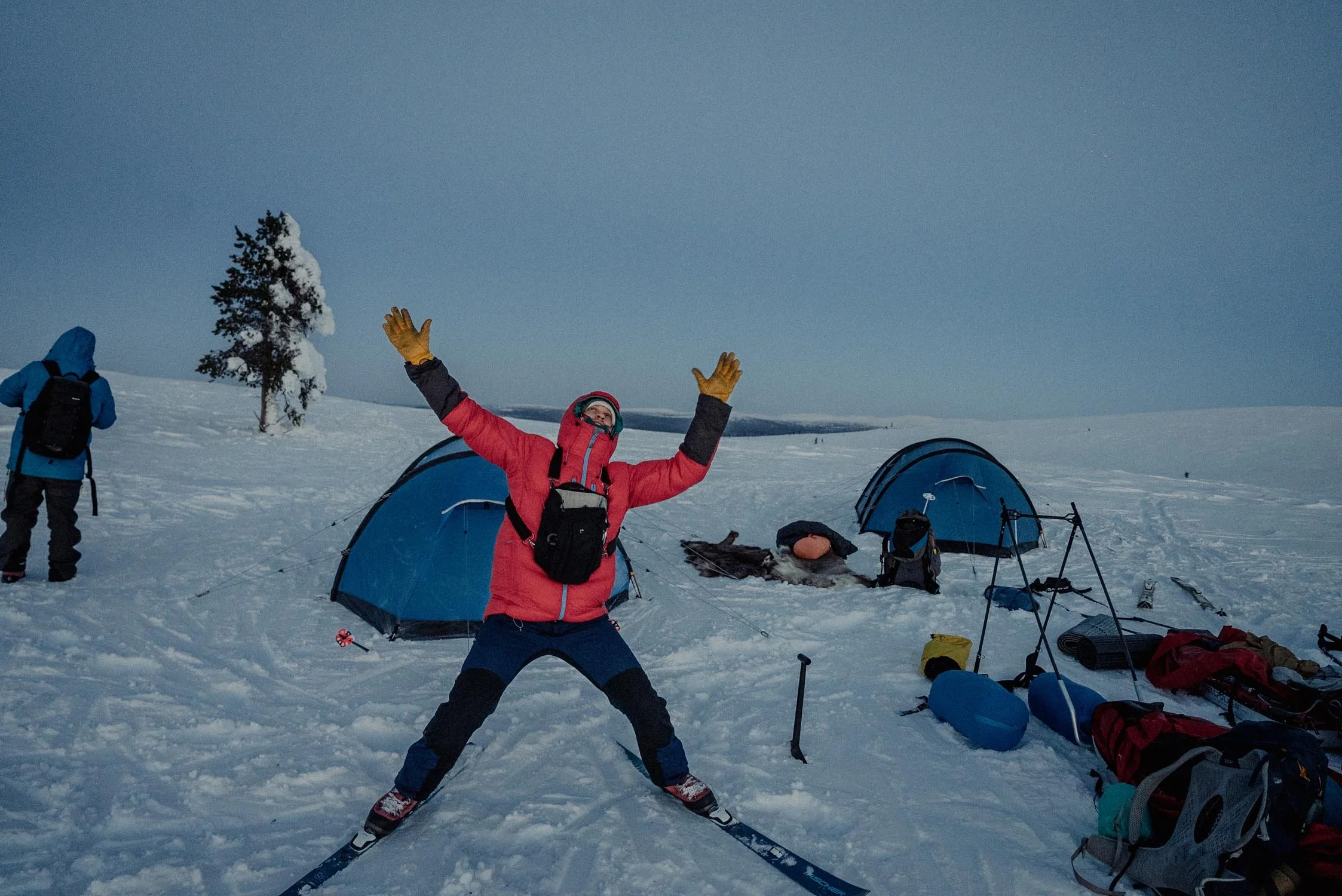 Person in a red jacket and yellow gloves crossing skis on snow, arms raised, with camping tents and gear nearby in a snowy landscape.