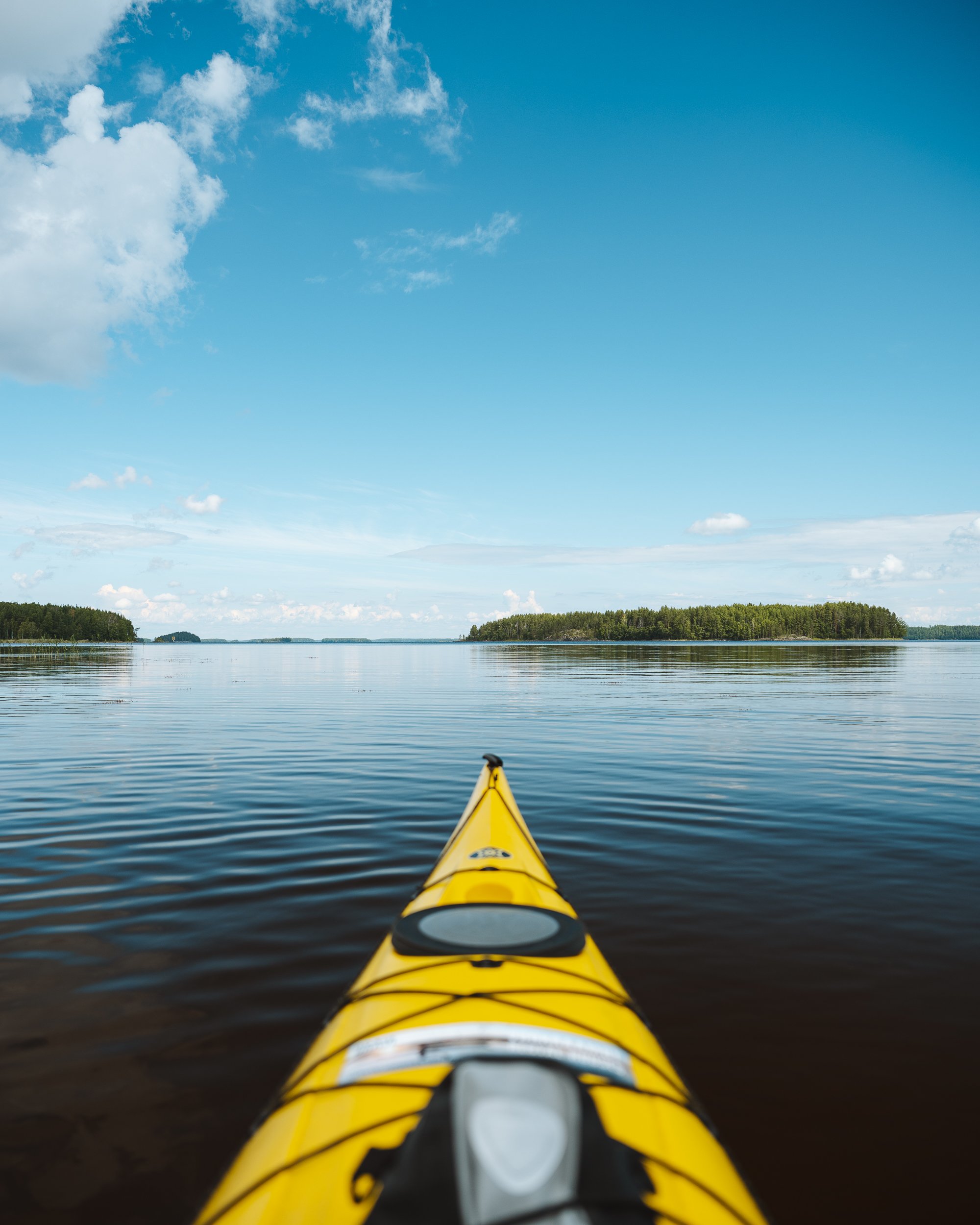 View from a kayak looking over the water to a distant tree-lined shoreline under a blue sky with scattered clouds.