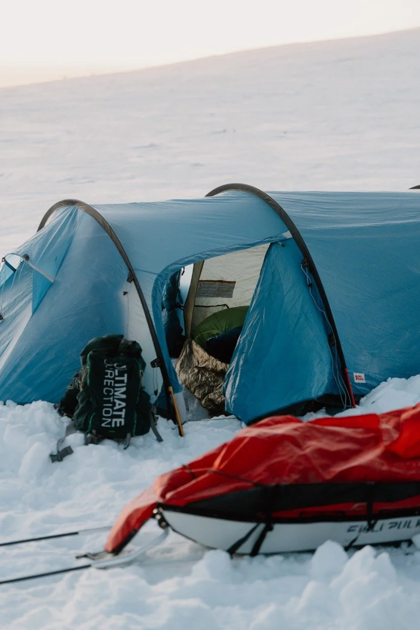 A blue camping tent set up in the snow with a backpack nearby and a red sled or kayak in the foreground, amid a snowy landscape.