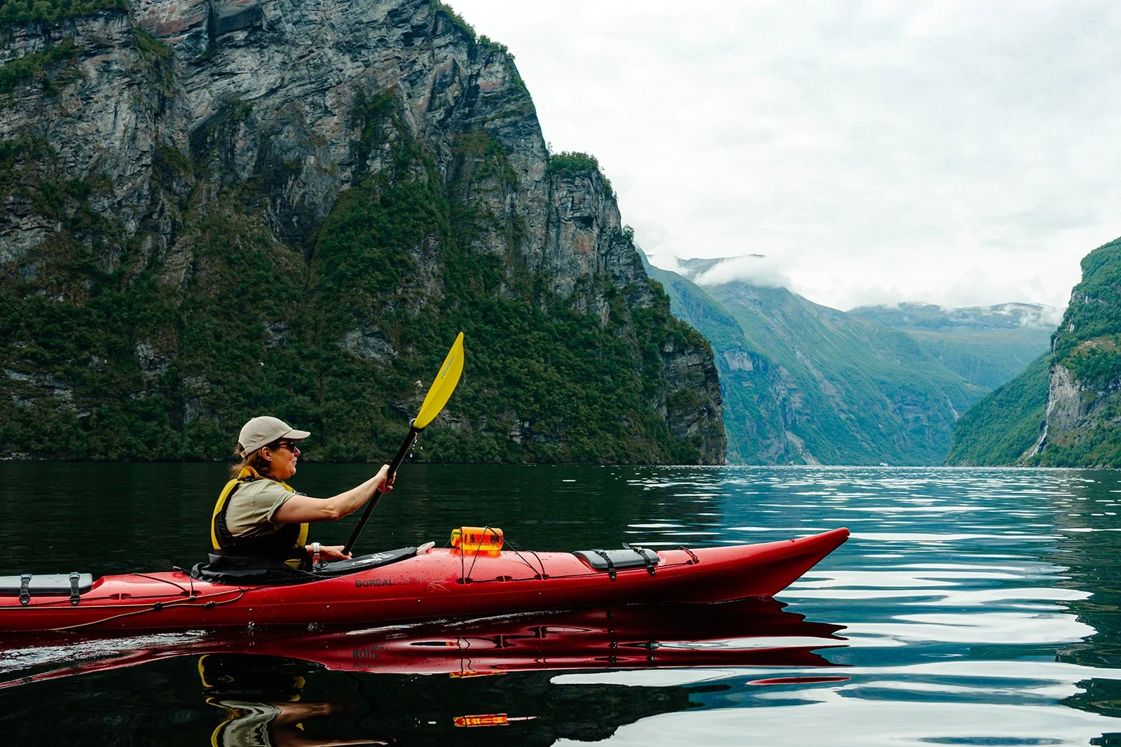 A person kayaking in a scenic Norwegian fjord with steep green cliffs on either side and cloudy skies overhead.