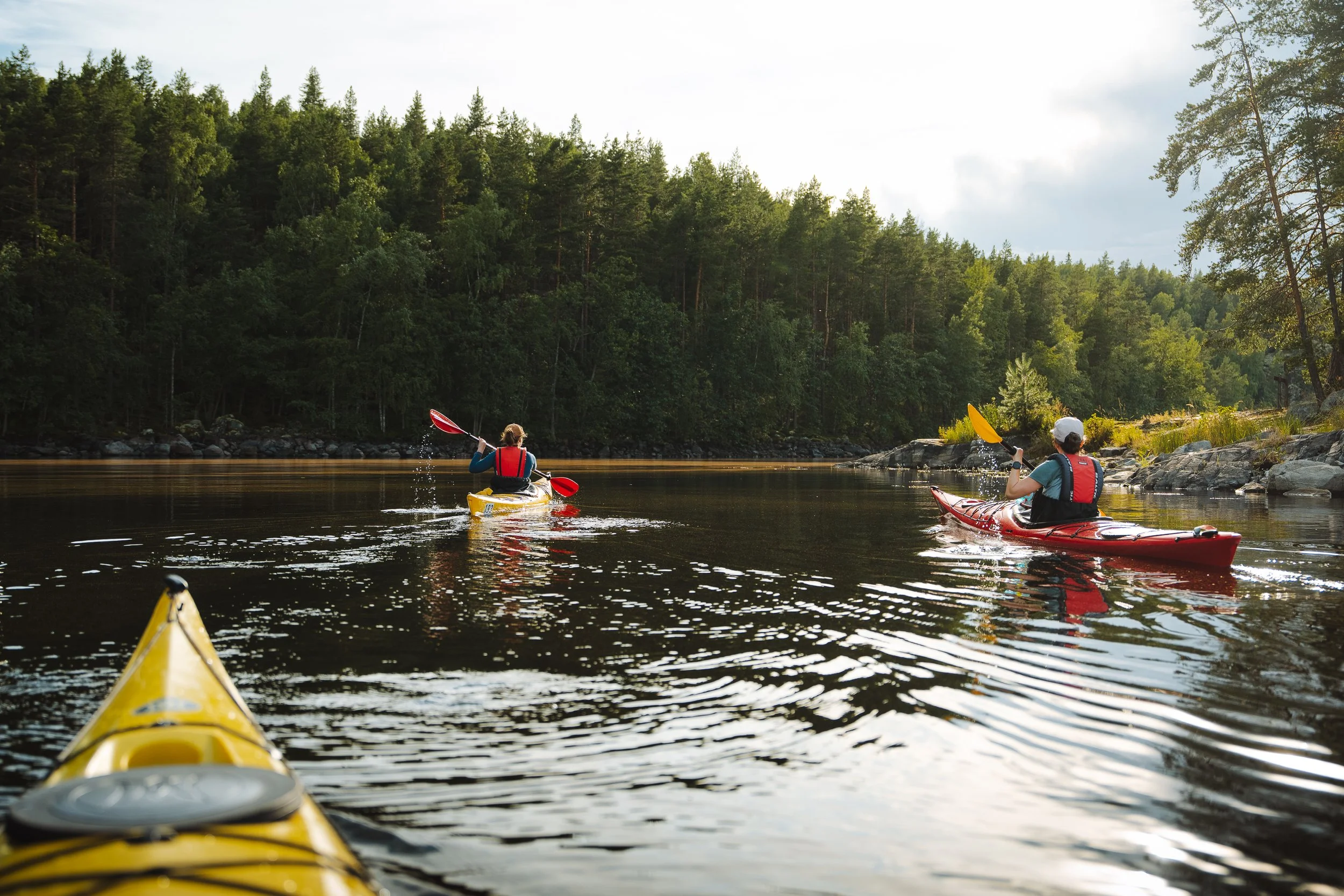 Two people kayaking on a calm river surrounded by dense green forest under a partly cloudy sky.