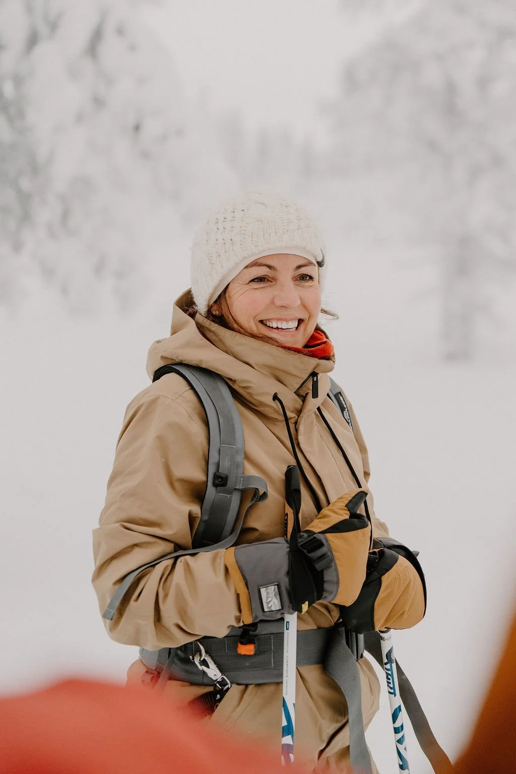 A woman in winter gear smiling outdoors in a snowy landscape, holding ski poles.