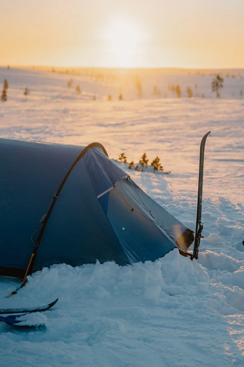 A blue tent set up in a snowy landscape during sunset, with snow-covered trees and horizon in the background.