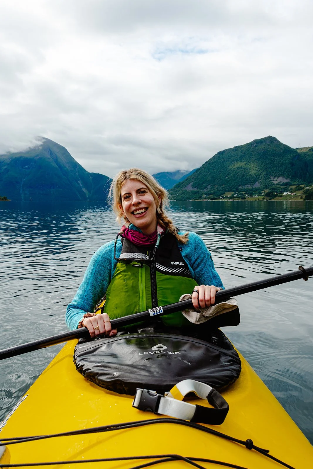 A woman with blonde hair smiling while kayaking on a lake surrounded by green mountains and cloudy sky.