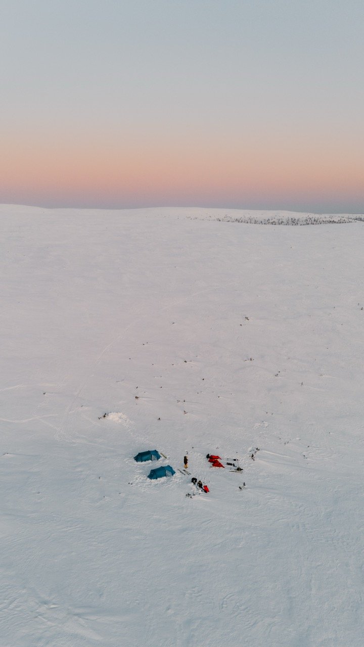 A winter landscape with a snow-covered plain and a few tents set up, with snowmobiles nearby and a person standing outside. The sky has a gradient of pink, purple, and blue as the sun sets or rises.