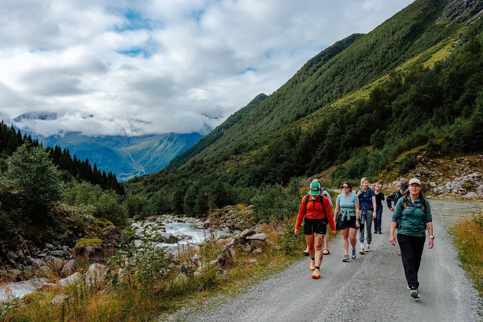 A group of seven hikers walking along a gravel trail in a lush green mountain valley. The trail is next to a flowing stream and surrounded by dense shrubs and trees. Tall mountains with partly cloudy skies are visible in the background.