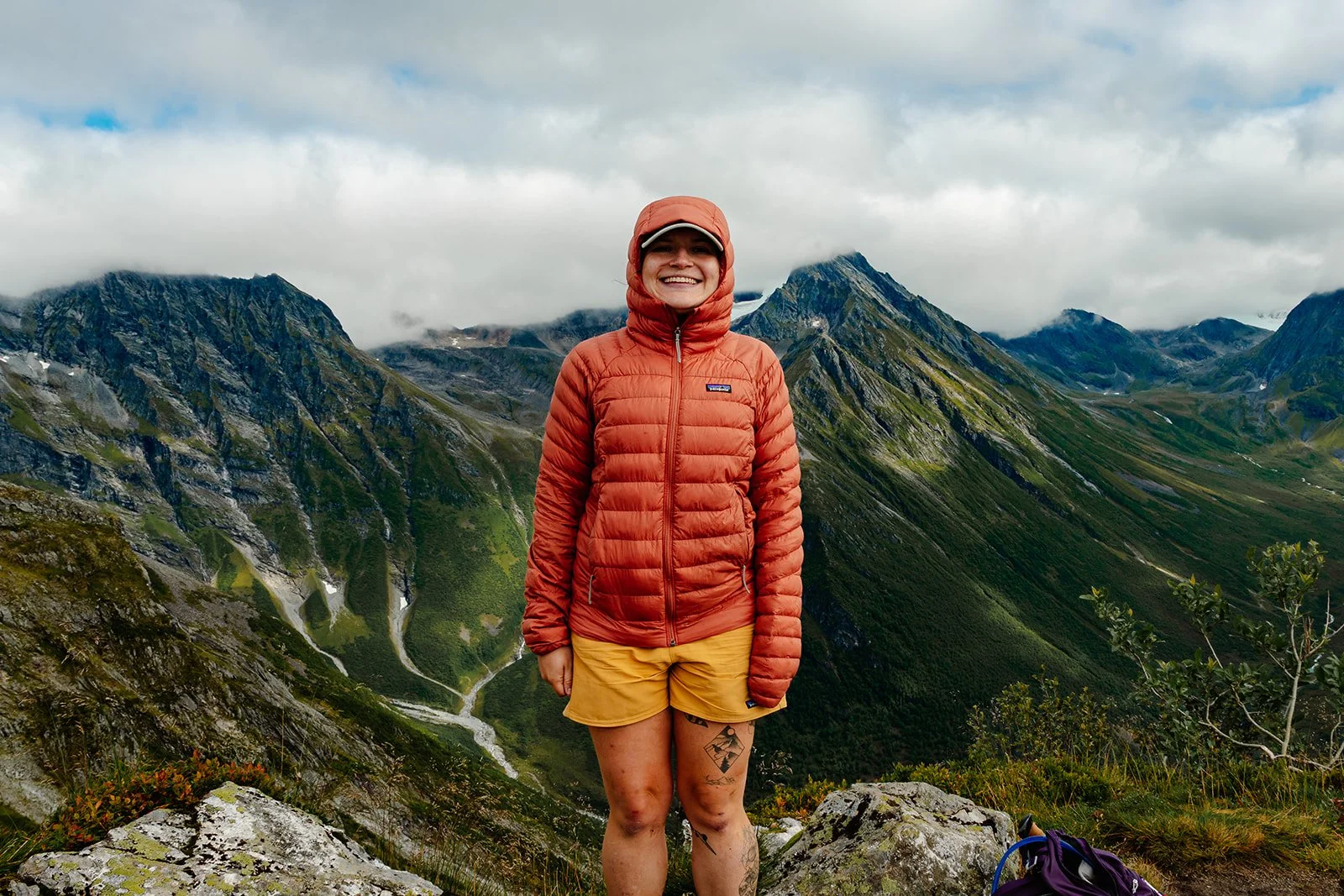 Woman hiking in a mountain landscape, wearing a red jacket, yellow shorts, and a beige cap, with mountains and clouds in the background.