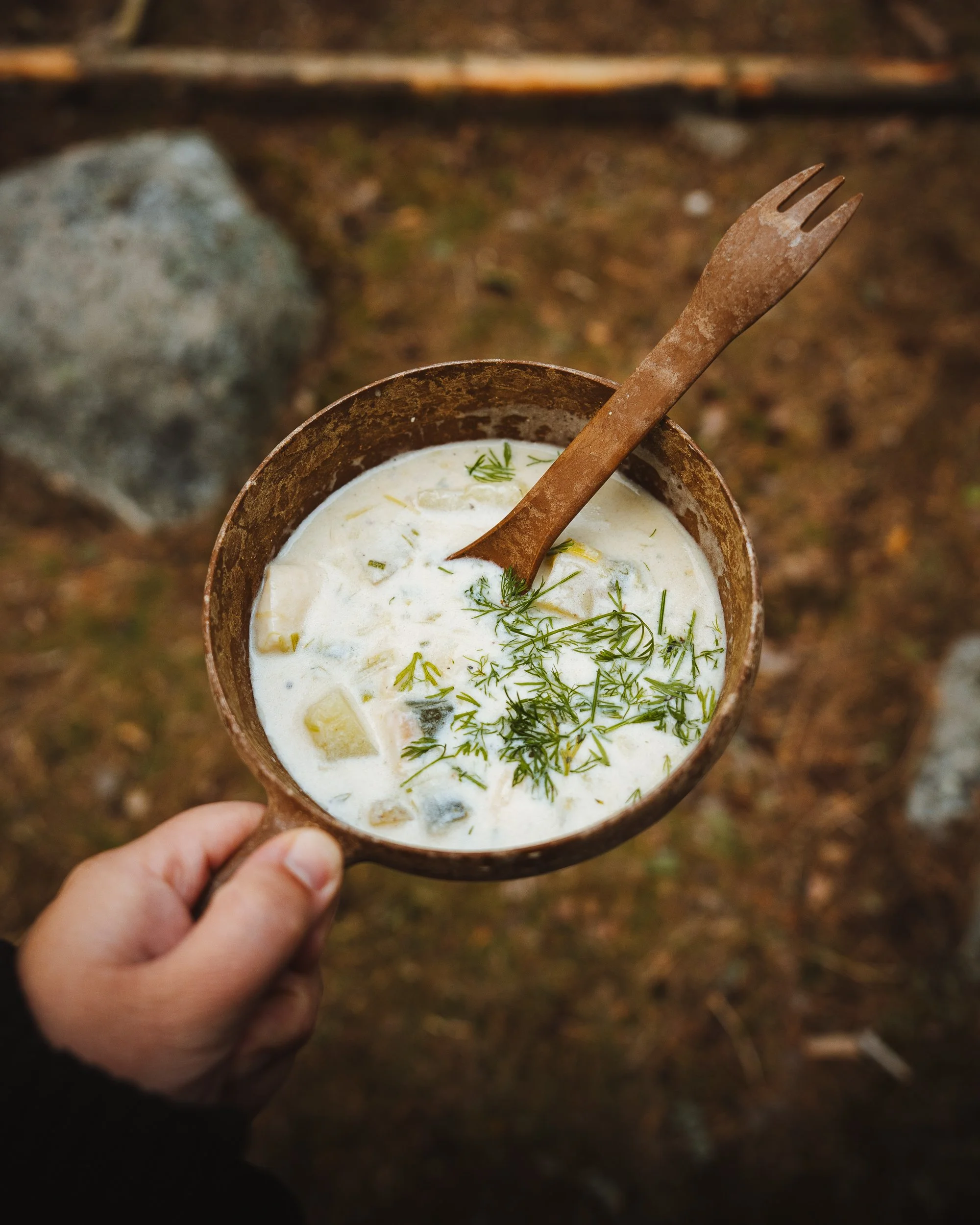 A hand holding a rustic metal cup with a creamy soup garnished with green herbs, with a rusty fork resting in the cup, outdoor background with woods and rocks.
