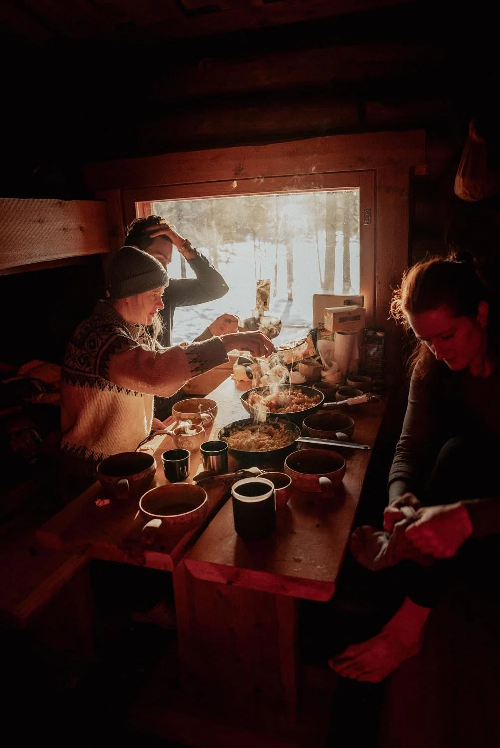 People having breakfast in a cozy cabin kitchen with sunlight streaming through a window, using bowls and mugs on a wooden table.