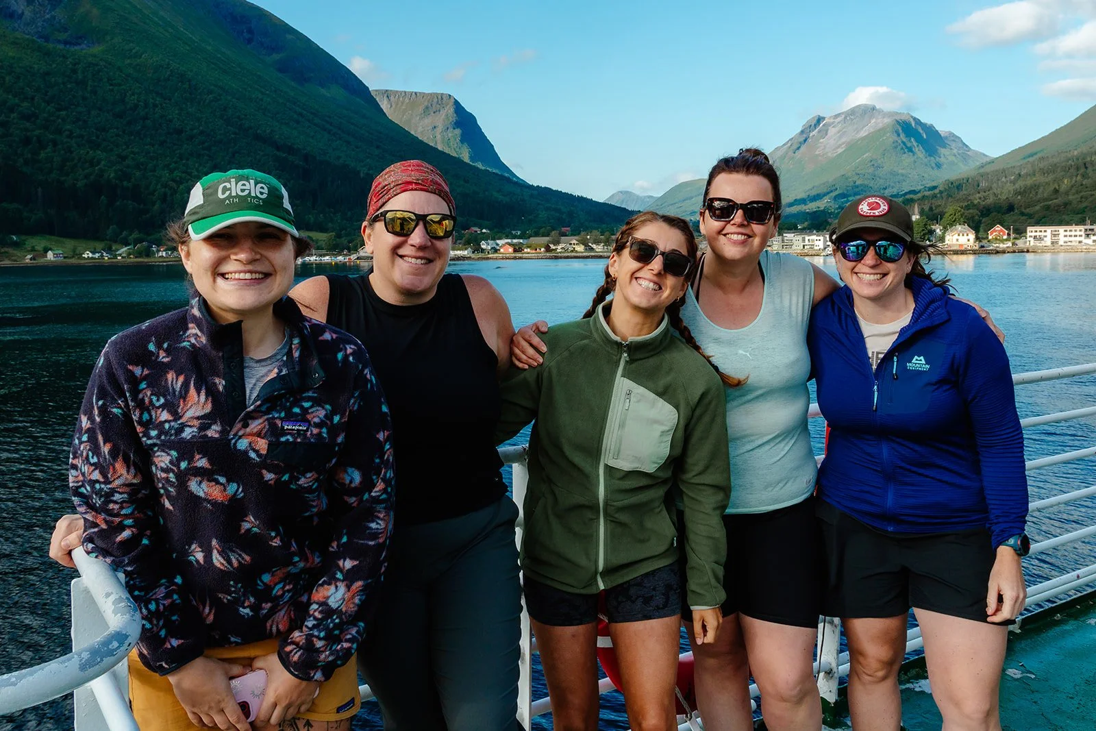 Five women smiling and posing together on a boat with a scenic backdrop of mountains, water, and a small town.