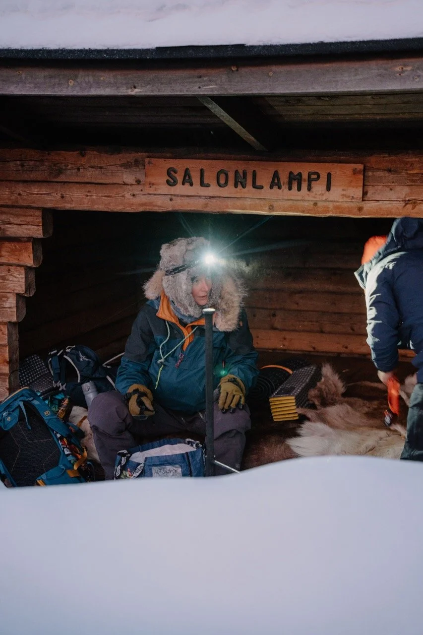 A person wearing winter gear and a furry hat, sitting inside a wooden shelter labeled 'SALONLAMP,' with outdoor gear around, and another person partially visible beside them.