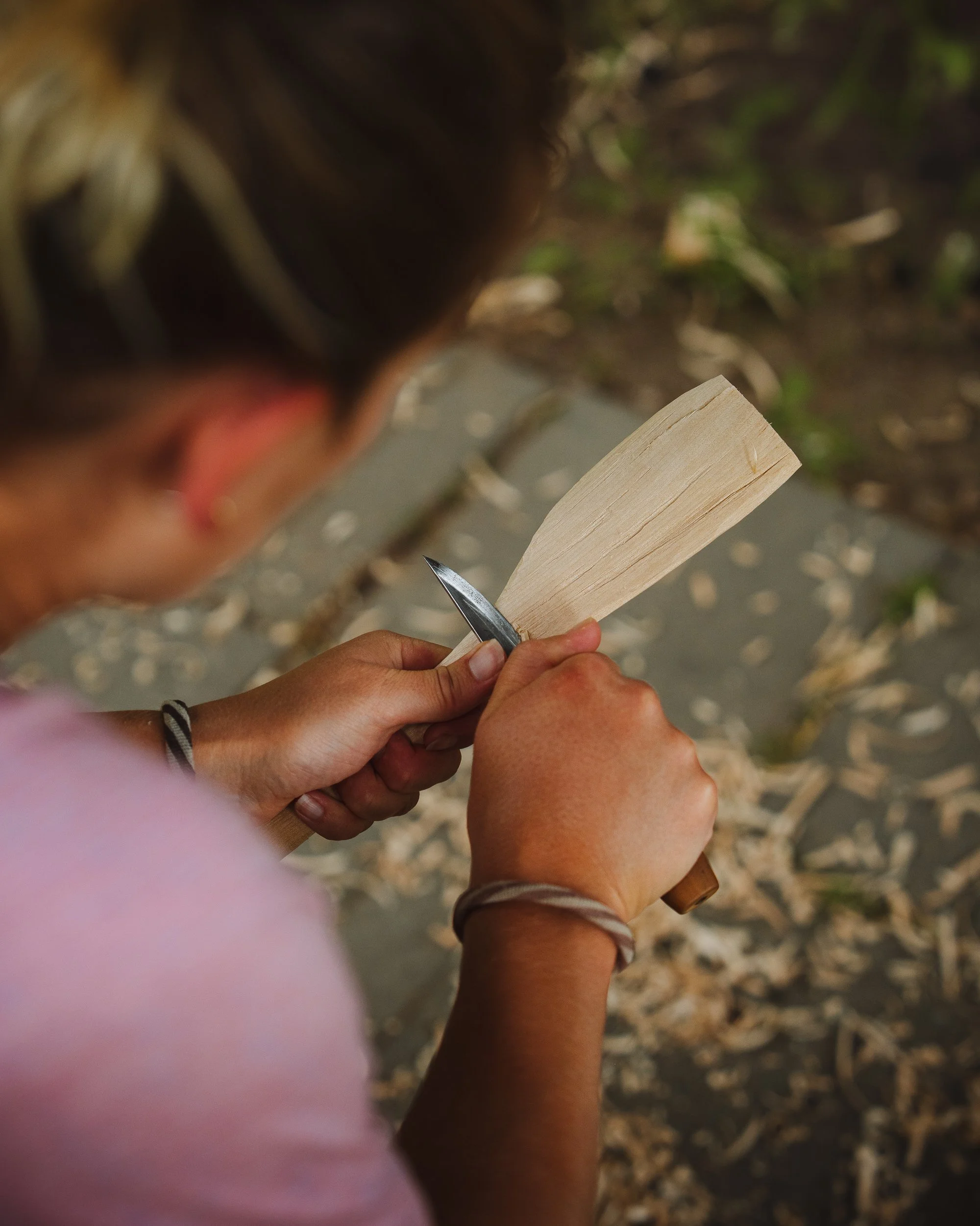 Close-up of hands sawing a light-colored piece of wood with a small hand saw, outdoors with blurred green foliage and ground.