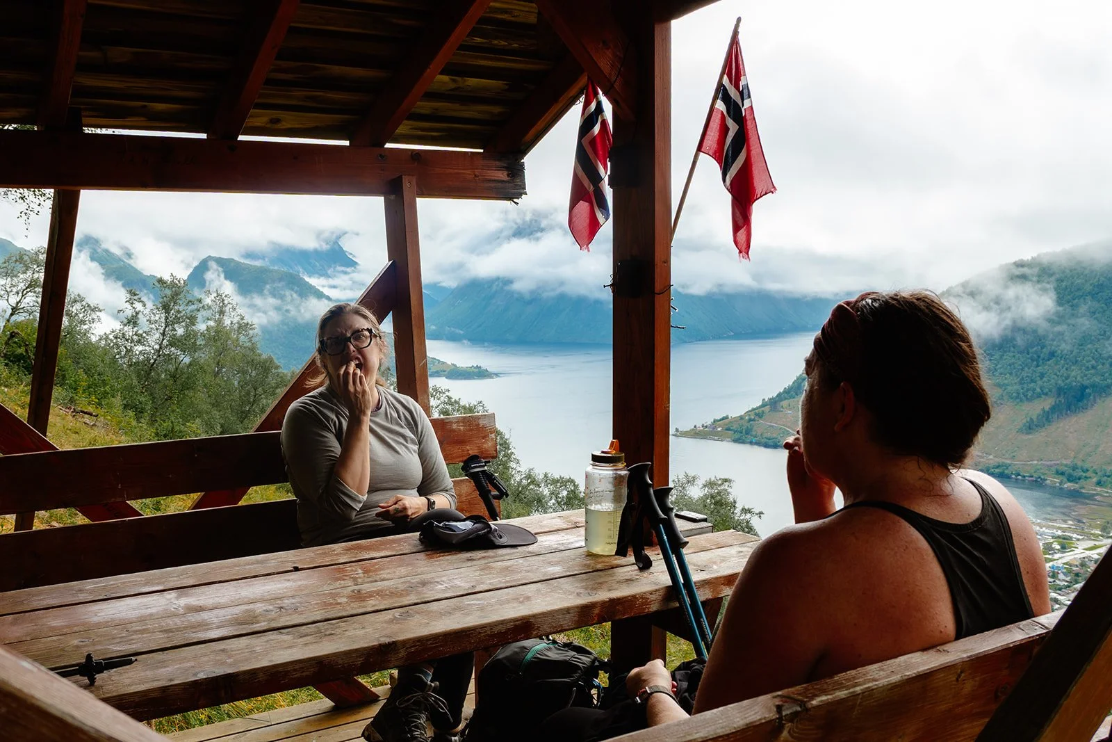 Two women sitting at a wooden table in a balcony with a scenic view of a lake, mountains, and clouds. One woman is biting her nails, and the other is facing her. There are water bottles, hiking gear, and national flags hanging from the balcony roof.
