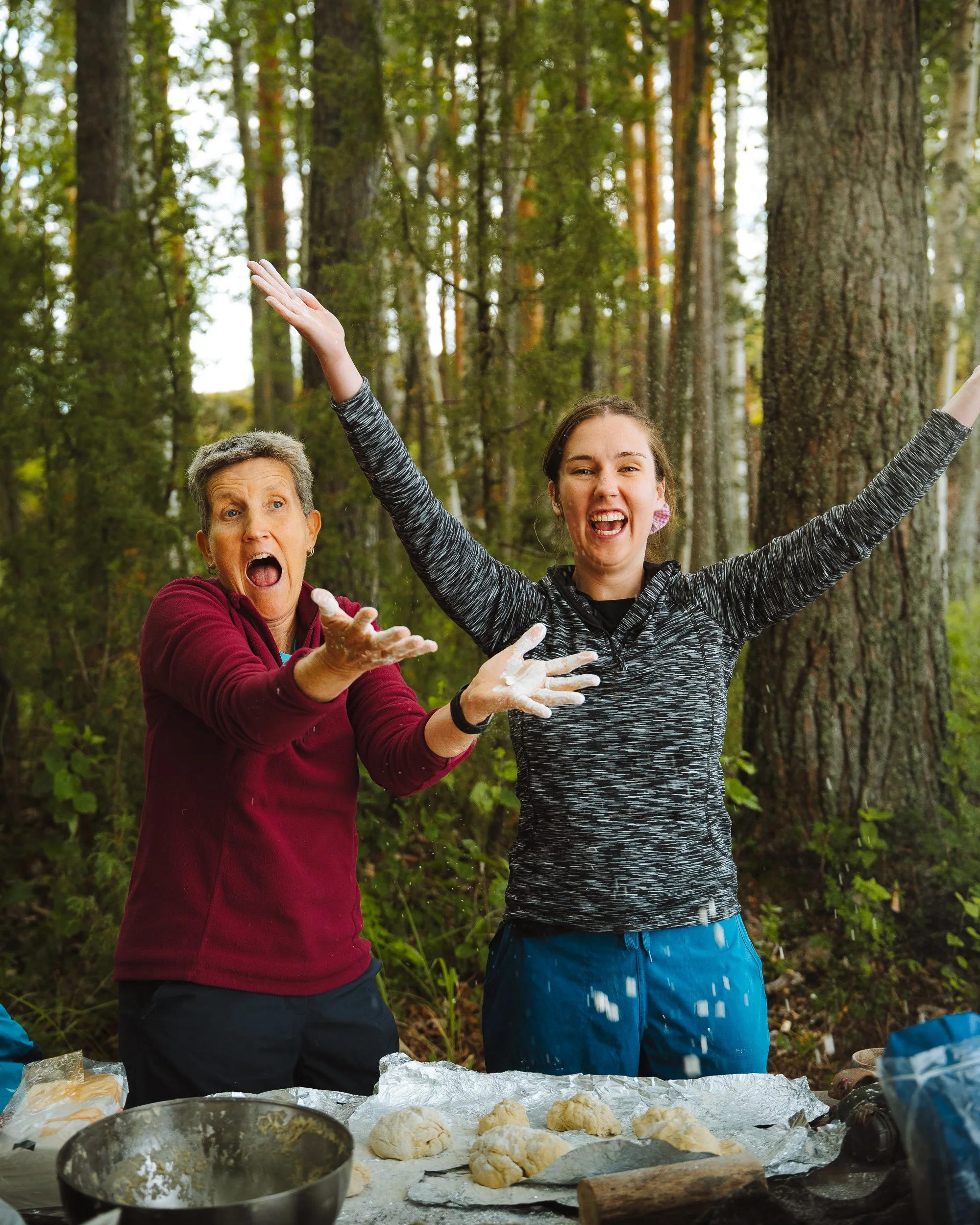 Two people, an older woman and a younger woman, preparing dough outdoors in a forest, with baking ingredients and dishes on a picnic table.