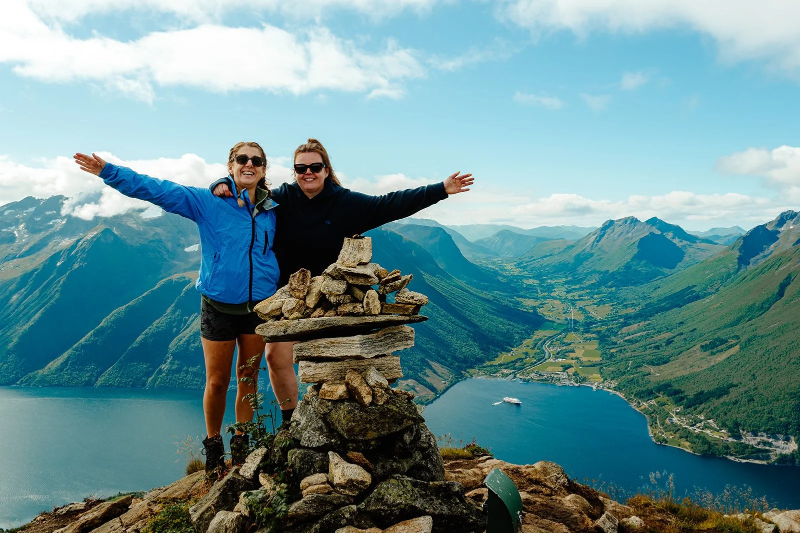 Two women smiling with arms outstretched on a mountain top In Norway, with a large body of water and green valleys surrounded by mountains in the background.