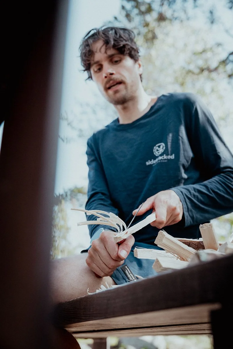 A man outdoors carving wood with a knife, focused on his work with wood shavings around him.