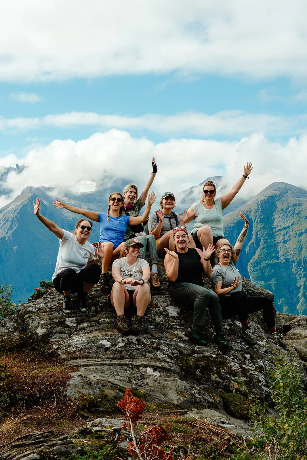 Group of nine women celebrating on top of a rocky mountain with a scenic mountain range and sky in the background in Norwegian fjords.