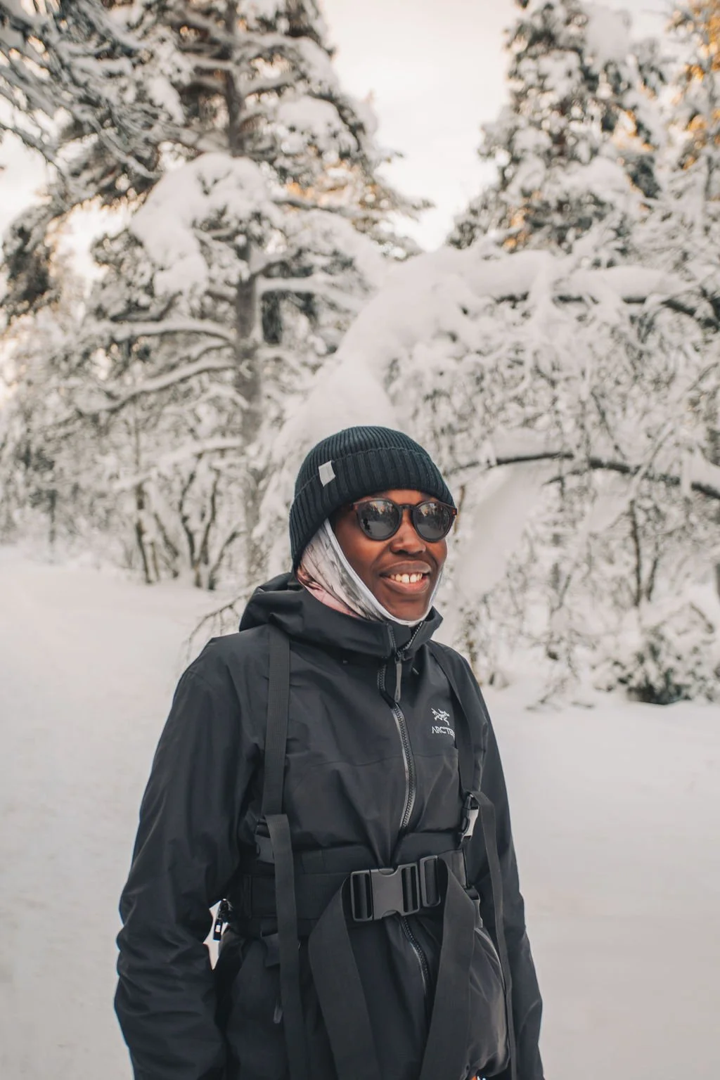 A smiling person wearing sunglasses, a black beanie, and a black jacket, standing in a snowy forest.