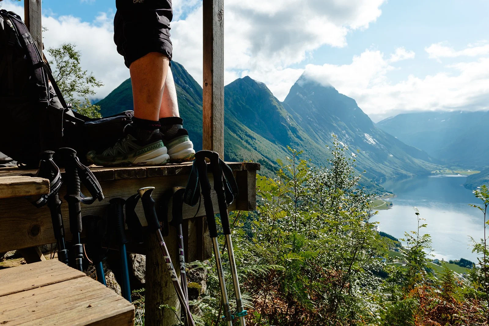 Hiking boots and trekking poles on a wooden platform overlooking a scenic mountain landscape with a lake and lush greenery.