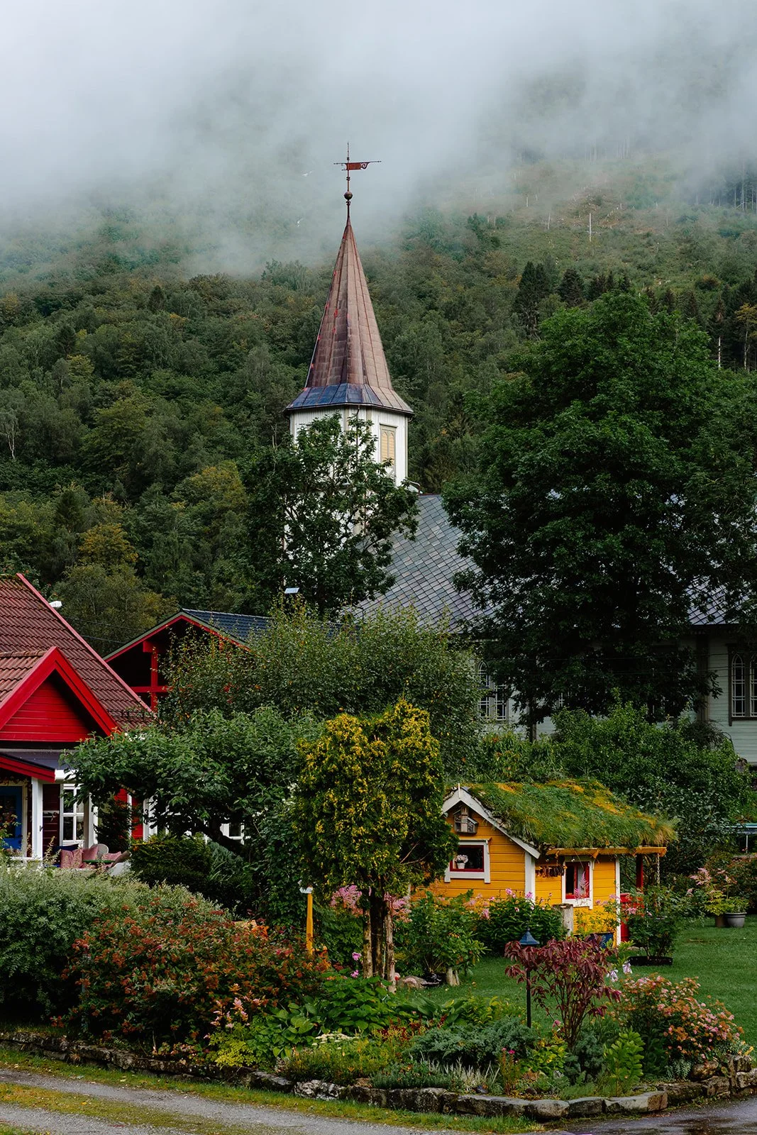 A scenic view of a church steeple with a weather vane, surrounded by lush green trees and colorful houses in a mountain village with fog-covered mountains in the background.