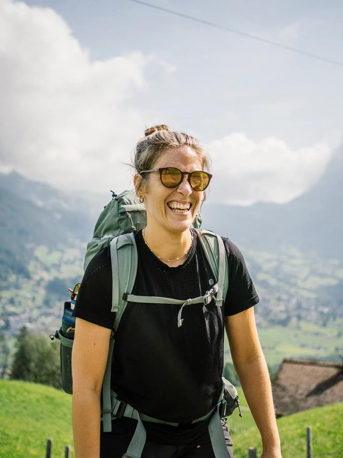 A woman with sunglasses smiling outdoors in a mountainous landscape, wearing a black t-shirt and carrying a backpack.
