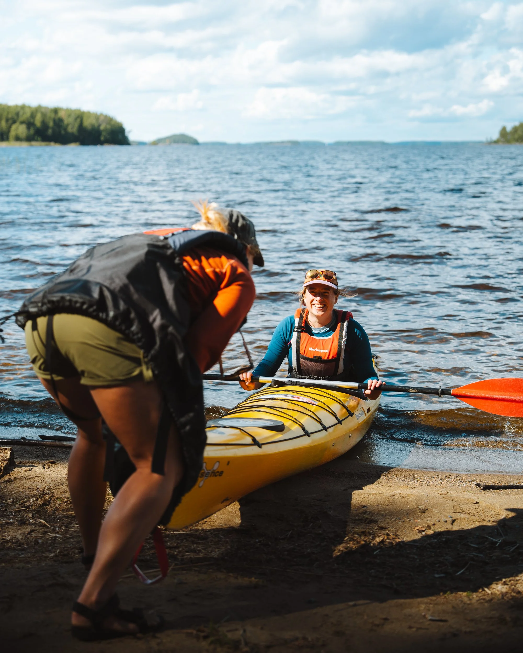Two people preparing a yellow kayak on the shore by a lake, with water and trees in the background, one of them smiling and the other bending over to adjust the kayak.