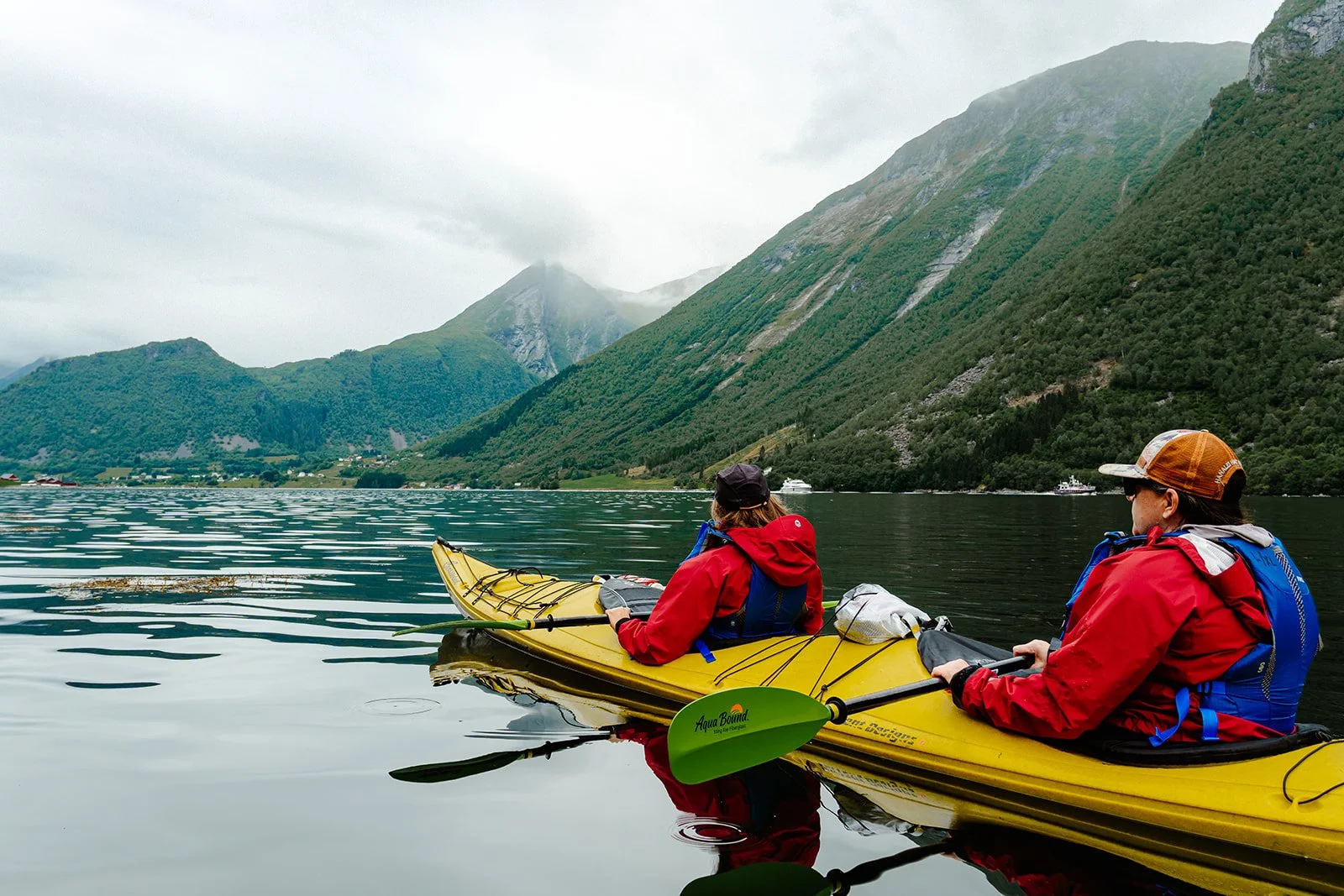 Two people in red jackets kayaking on a calm body of water surrounded by green mountains with a cloudy sky in Norway.