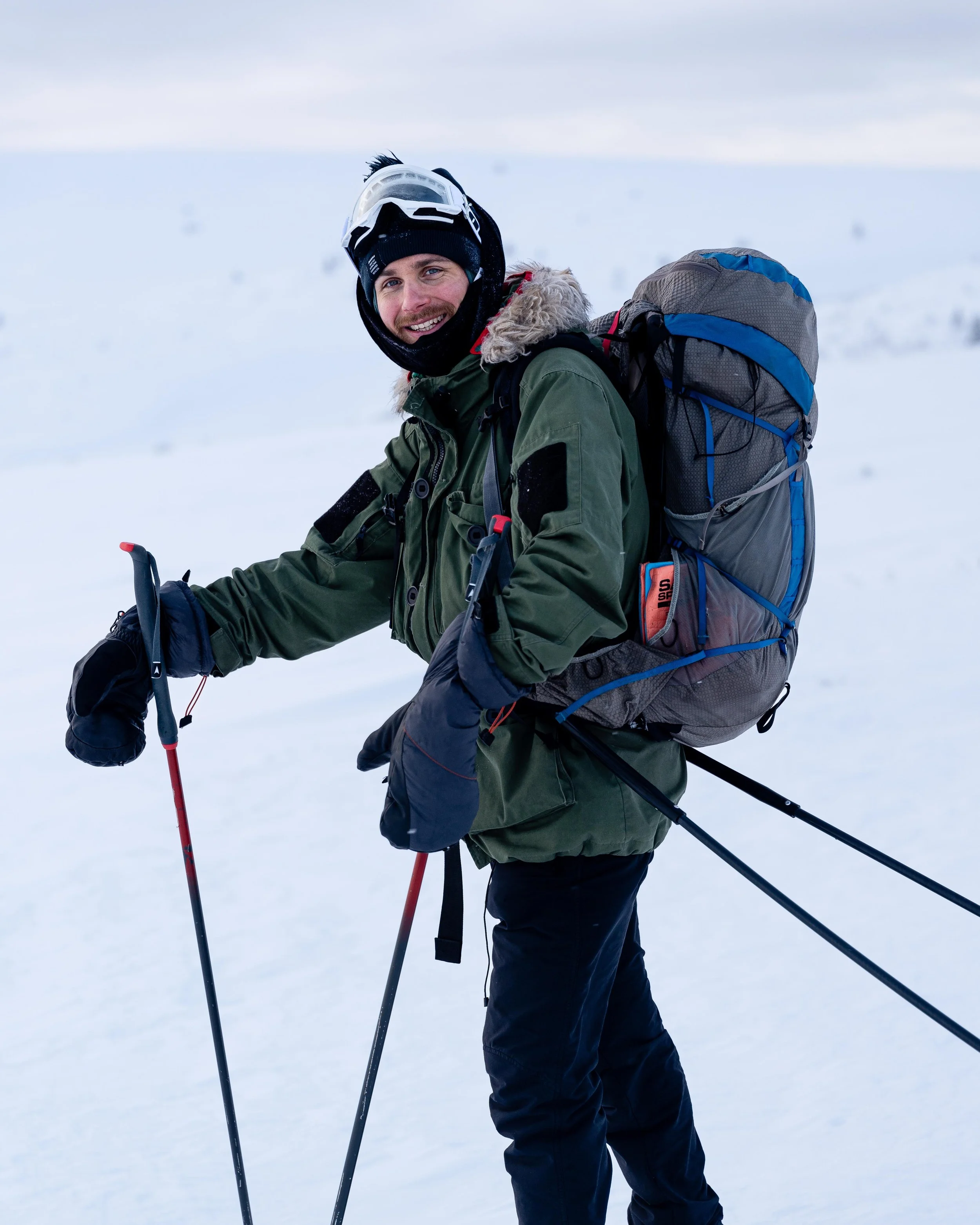 A man in winter outdoor gear smiling while standing on snow, holding ski poles, with a large backpack, wearing a helmet and goggles, in a snowy landscape.