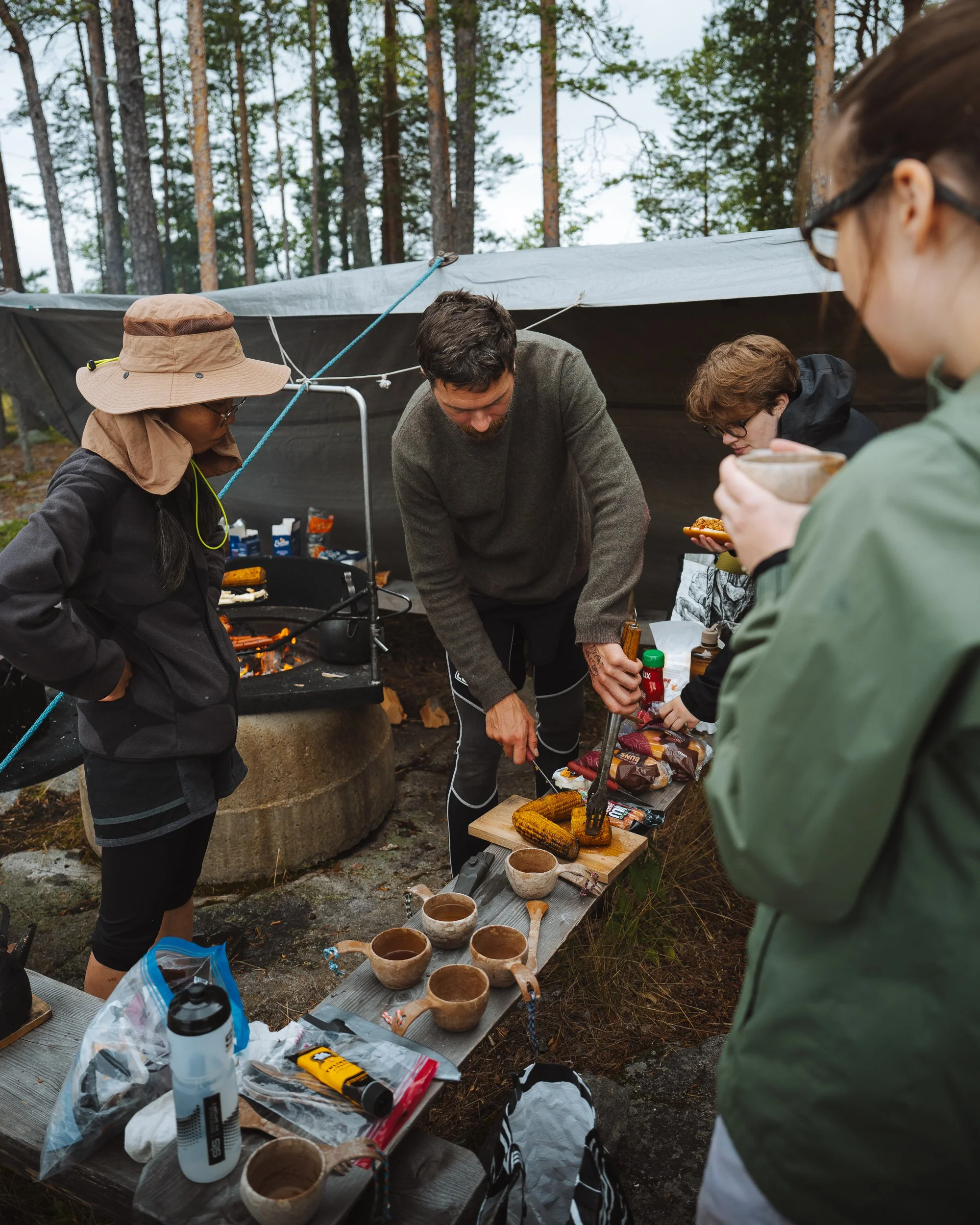 A group of people outdoors at a campsite, preparing food and drinks near a table with pottery and utensils, and a tent set up in a forested area.