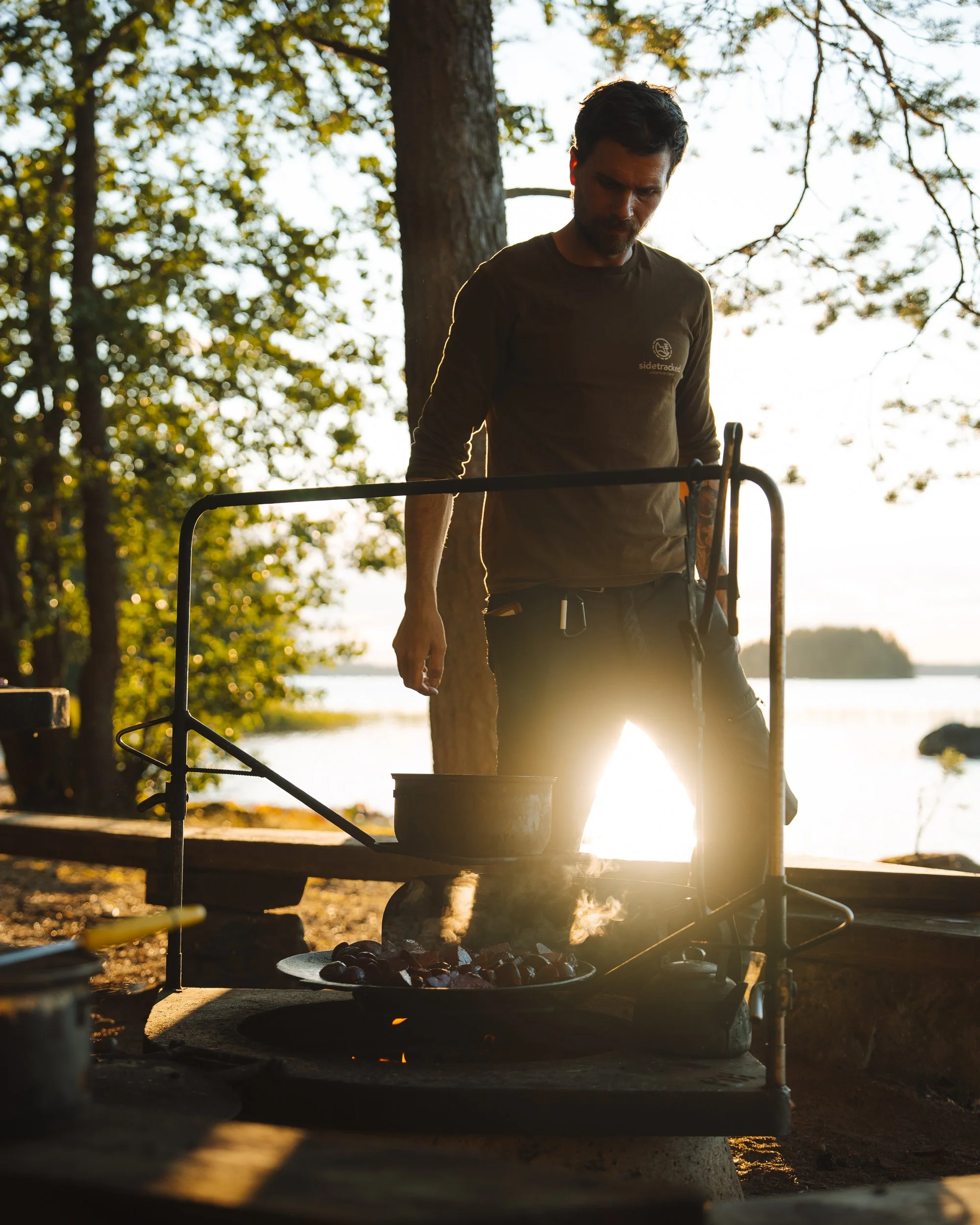 A man standing outdoors near a lake, cooking food over a small grill, backlit by the setting sun with trees in the background.
