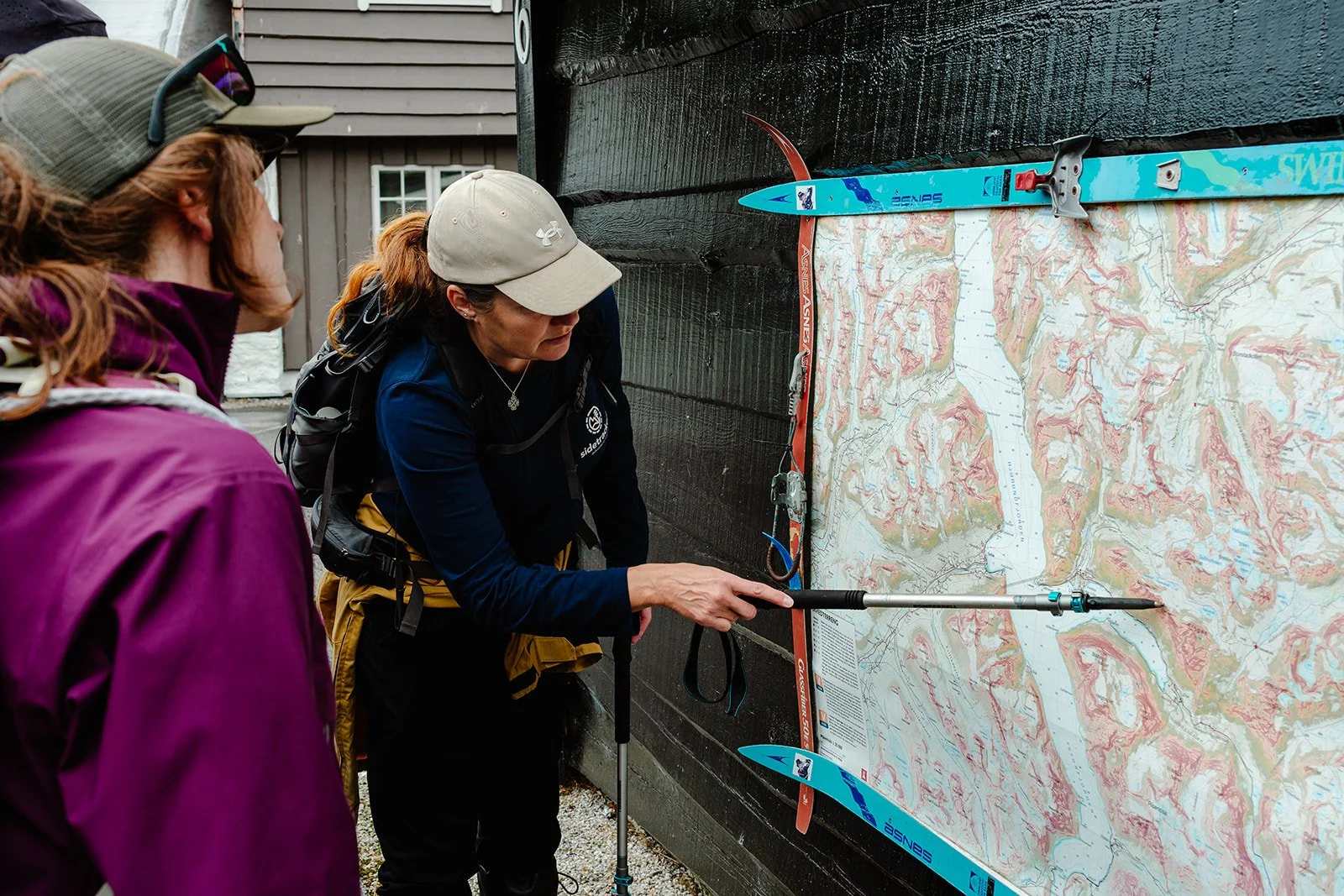 Two women standing outdoors near a topographical map on a black wall. One woman is using a trekking pole to point at the map, while the other watches. The map is attached to the wall with two alpine skis.