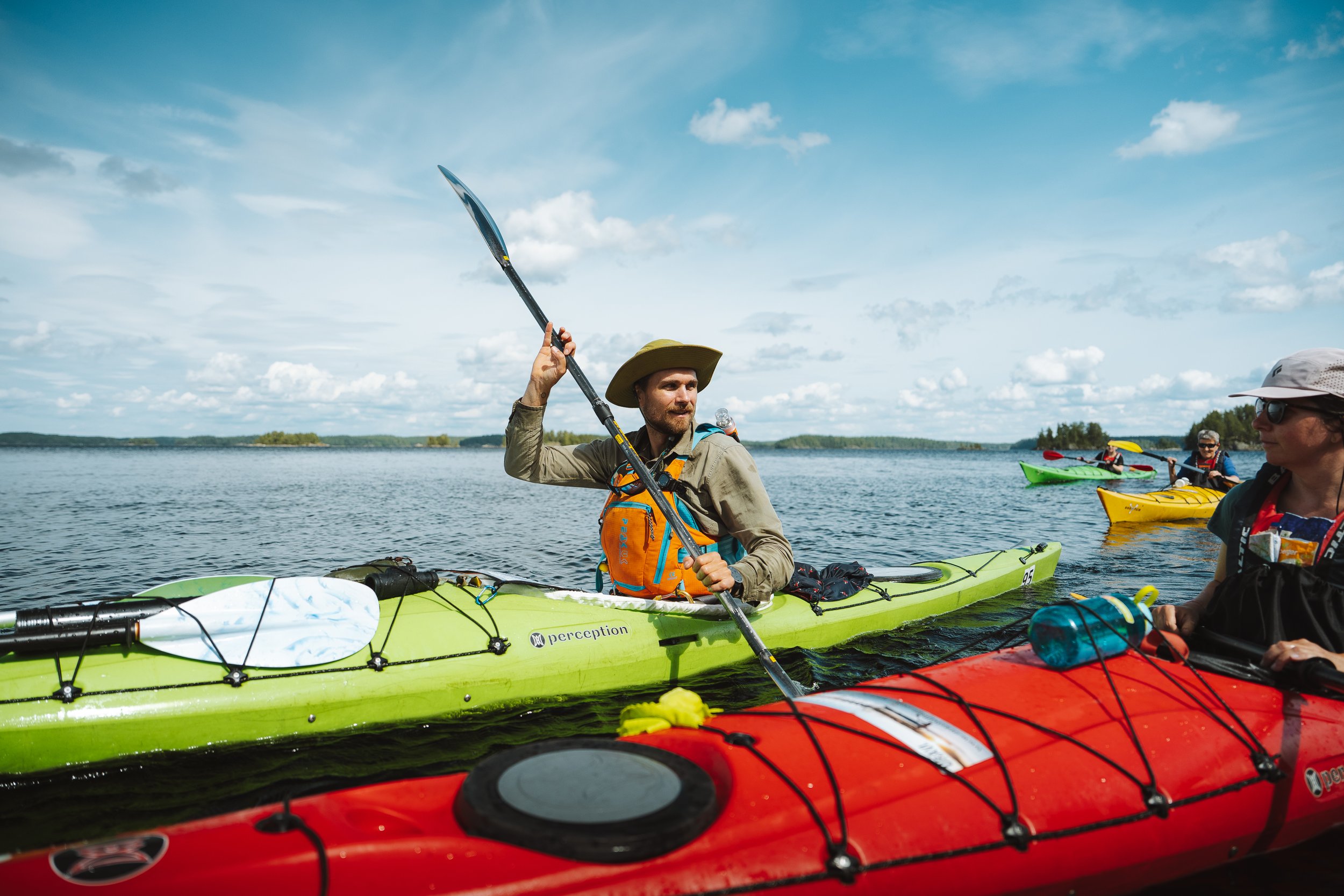 People kayaking on a lake under a blue sky with clouds, wearing hats and life jackets.