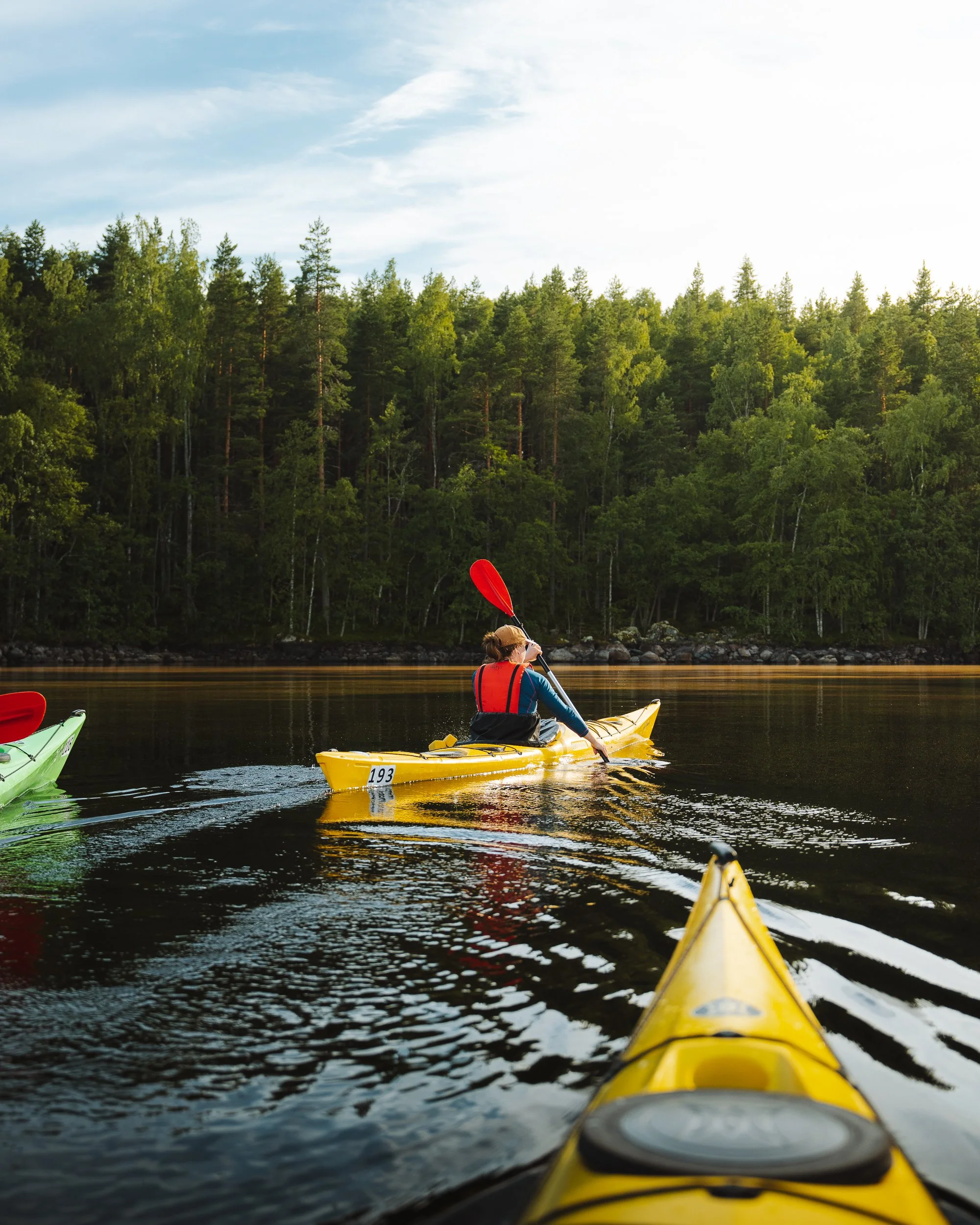 View from a yellow kayak as it floats on a calm river, with a person in a red life jacket and brown hat paddling while holding a red paddle. Dense green forest and a partly cloudy sky are in the background.