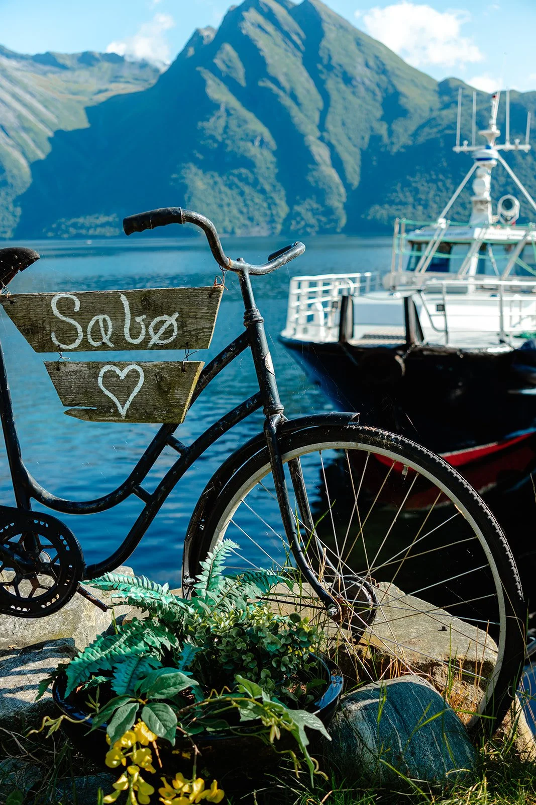 A black bicycle with a wooden sign that says "Saló" and a heart symbol attached to it, parked near a boat on a body of water with mountains in the background.